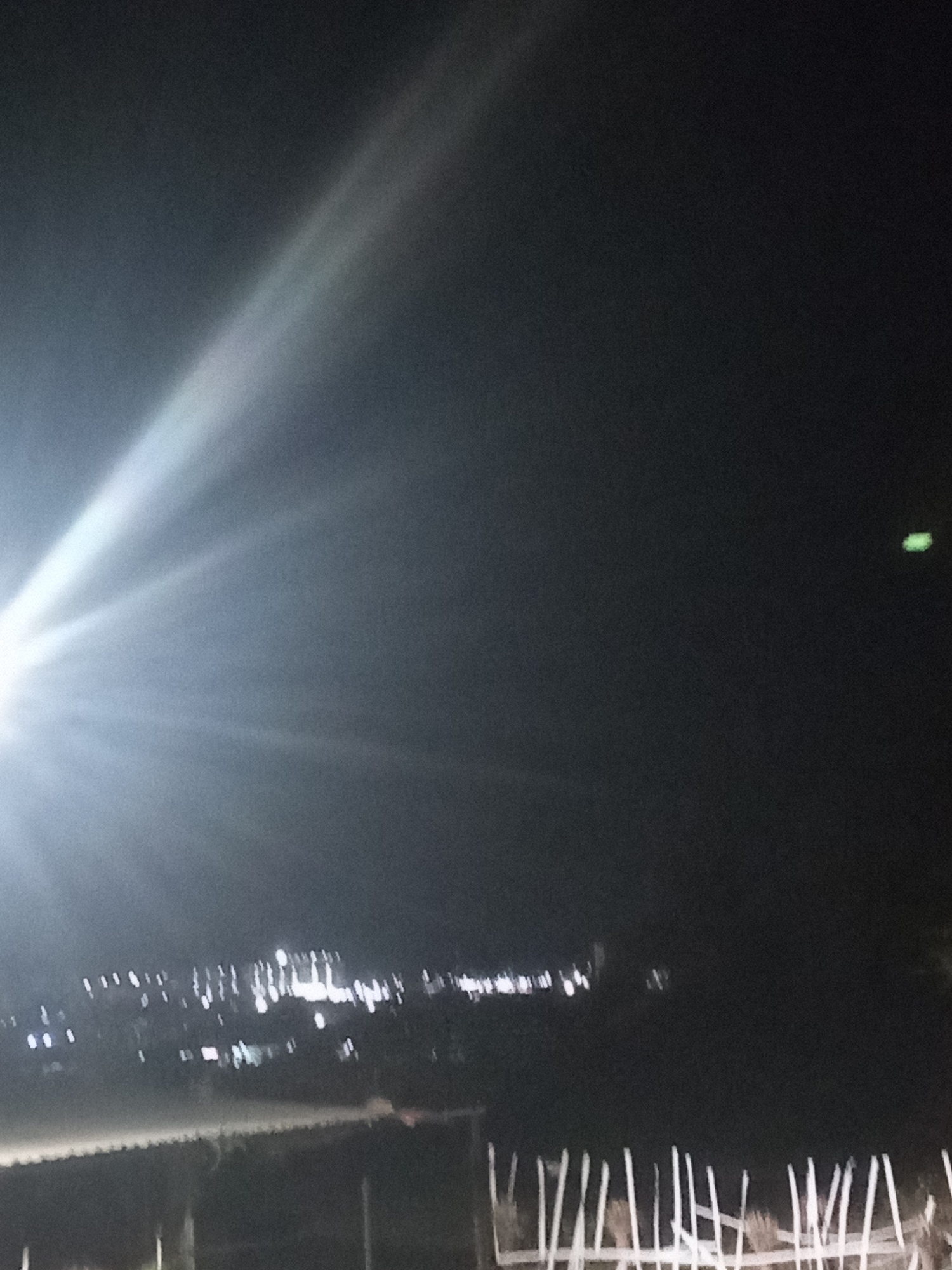 A night shot with extreme light sources. The horizon appears to have city lights, suggesting a populated area. There is a fence-like structure in the foreground and a light glare dominating the left side of the frame. No people are visible. Given the location Juan Griego, Venezuela, the city lights on the horizon are likely within the city limits, as this is a coastal location. There is also a bright green light on the right-hand side of the frame.