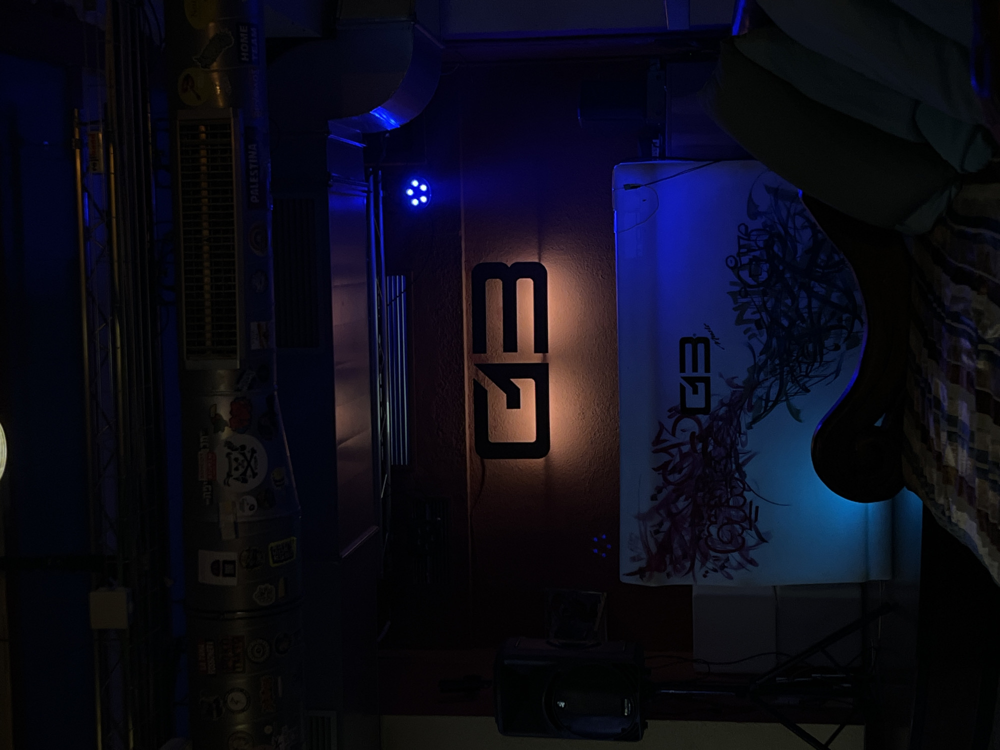 The scene is a dimly lit interior space, likely a bar or nightclub in Barcelona, Spain, based on the signage and ambiance. Prominently displayed is a stylized "13" logo, illuminated with warm light against a dark wall. To the right of the logo is a vertically oriented canvas or board with graffiti-style art, lit with blue light. A sound system, including a speaker and a small glass on top of it, sits below the wall art. To the left of the "13" logo, a vertical air duct or pipe is covered in stickers. A blue spotlight is directed towards the wall, adding to the ambient lighting. On the right side of the image, a draped object adds to the complexity of the scene. The overall impression is that of a contemporary urban environment with a vibrant nightlife.