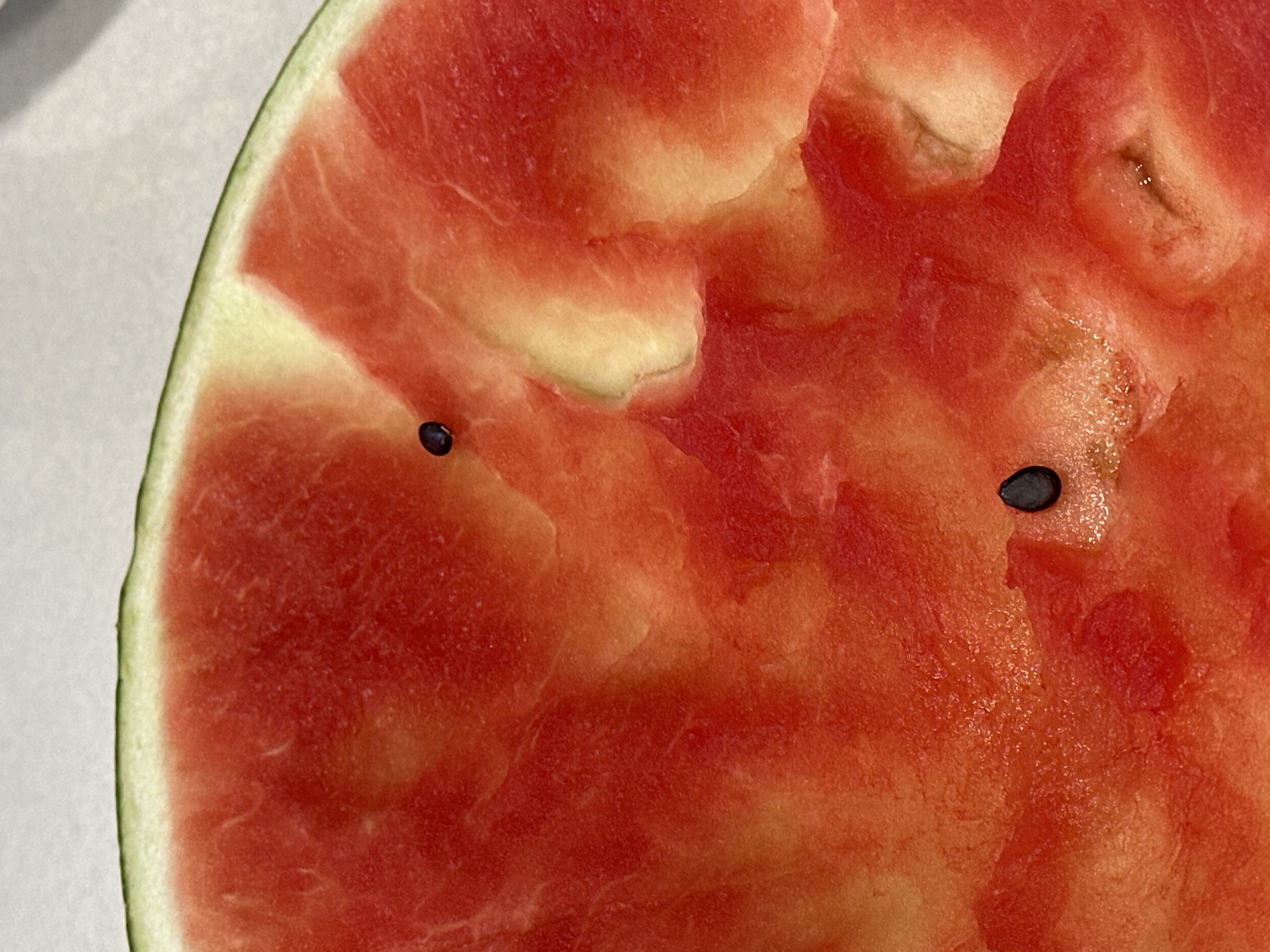 The image is a close-up of a slice of watermelon. The flesh of the watermelon is a vibrant red with some lighter yellow-white areas. There are three visible black seeds embedded in the flesh. The rind of the watermelon is green and light green. The background is a textured, light-colored surface, possibly a table. The image is taken from a slightly elevated angle, looking down into the slice of watermelon. There are no people or animals present in the image. The location in the city of Nevinnomyssk, Russia, is not discernible from the image itself.