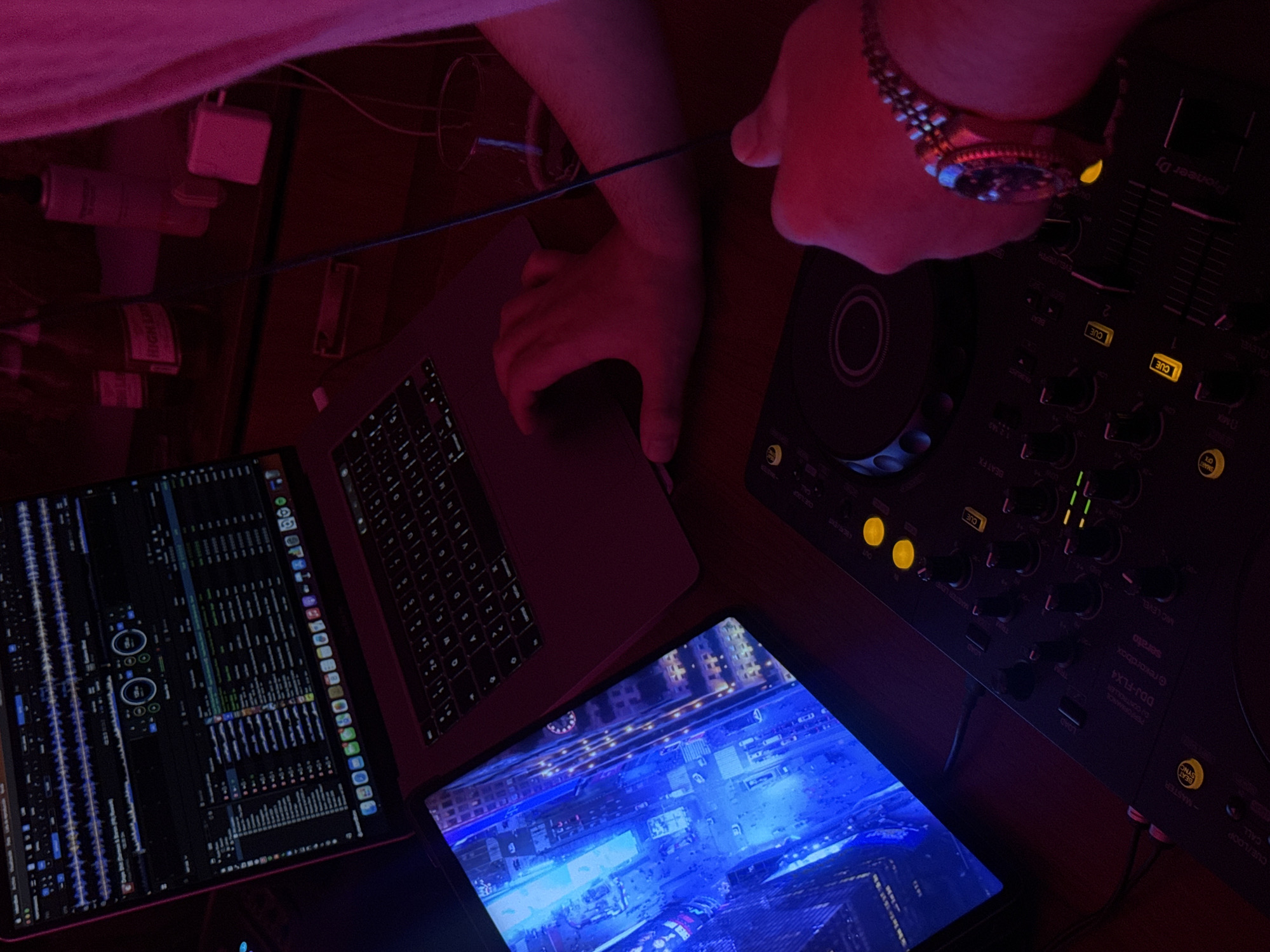The image shows a DJ setup under red and blue lighting. The setup includes a laptop, two tablets, and a Pioneer DJ DDJ-FLX4 controller. One tablet displays a waveform audio interface. One hand is operating the laptop, and another hand, with a watch, is poised over the DJ controller. There are a couple of adapters and cables scattered on the desk, along with a half-visible object in the upper left. The environment suggests an indoor setting, possibly a home studio. Location: The reference to Puebla, Mexico, is not visually evident in the image.