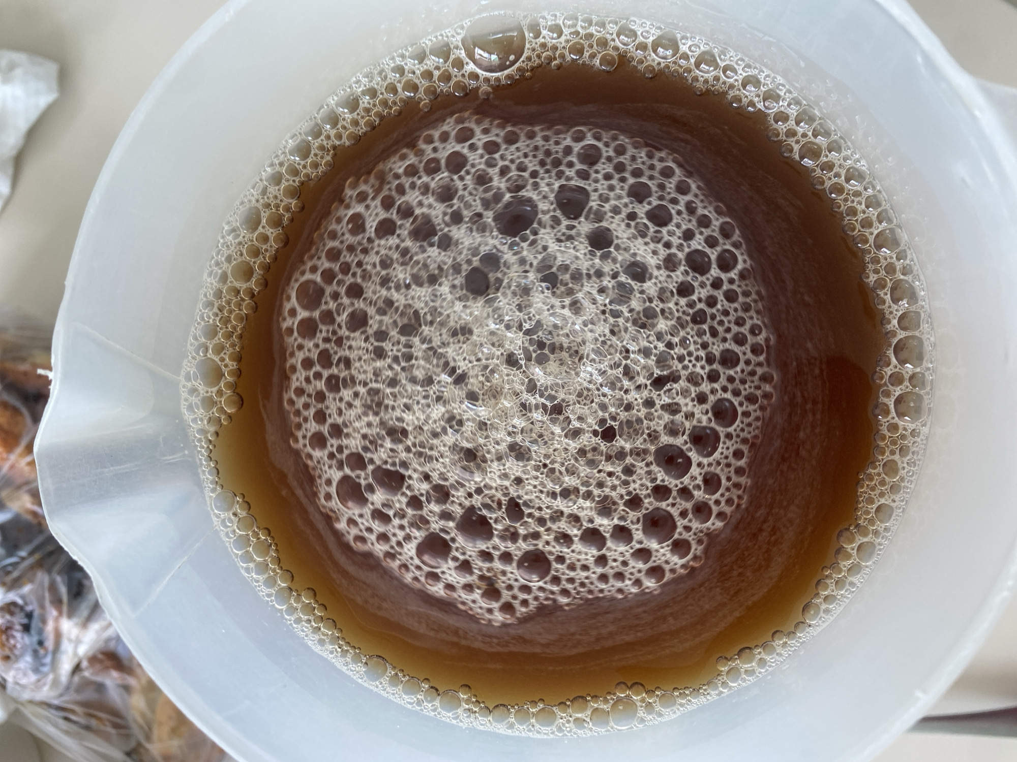 An overhead shot captures a dark amber or brown liquid contained within a white, translucent, possibly plastic, circular vessel, resembling a measuring cup or beaker. The liquid's entire surface is covered by a dense layer of off-white to light brown foam, consisting of numerous bubbles of varying sizes.
The bubbles form an intricate pattern: a central area features a tight cluster of smaller, very dense bubbles, which gradually transition to larger, more widely spaced bubbles towards the edges. A distinct ring of smaller, uniform bubbles adheres to the inner wall of the container where the liquid meets the side. Many bubbles show light reflections on their rounded surfaces, indicating a glossy or wet texture.
The container itself appears functional, with slightly irregular edges typical of molded plastic. In the blurred background on the left, hints of a lighter surface and some indistinct, brown-toned objects, possibly items wrapped in clear plastic, suggest an indoor setting like a kitchen or a workspace in San Bernardino Tlaxcalancingo, Mexico. No specific activity is actively depicted, but the presence of foam implies the liquid has recently been agitated, mixed, poured, or is undergoing a process such as fermentation or brewing. The lighting is bright and even, possibly natural daylight. No visible text or markings are present on the container or liquid.