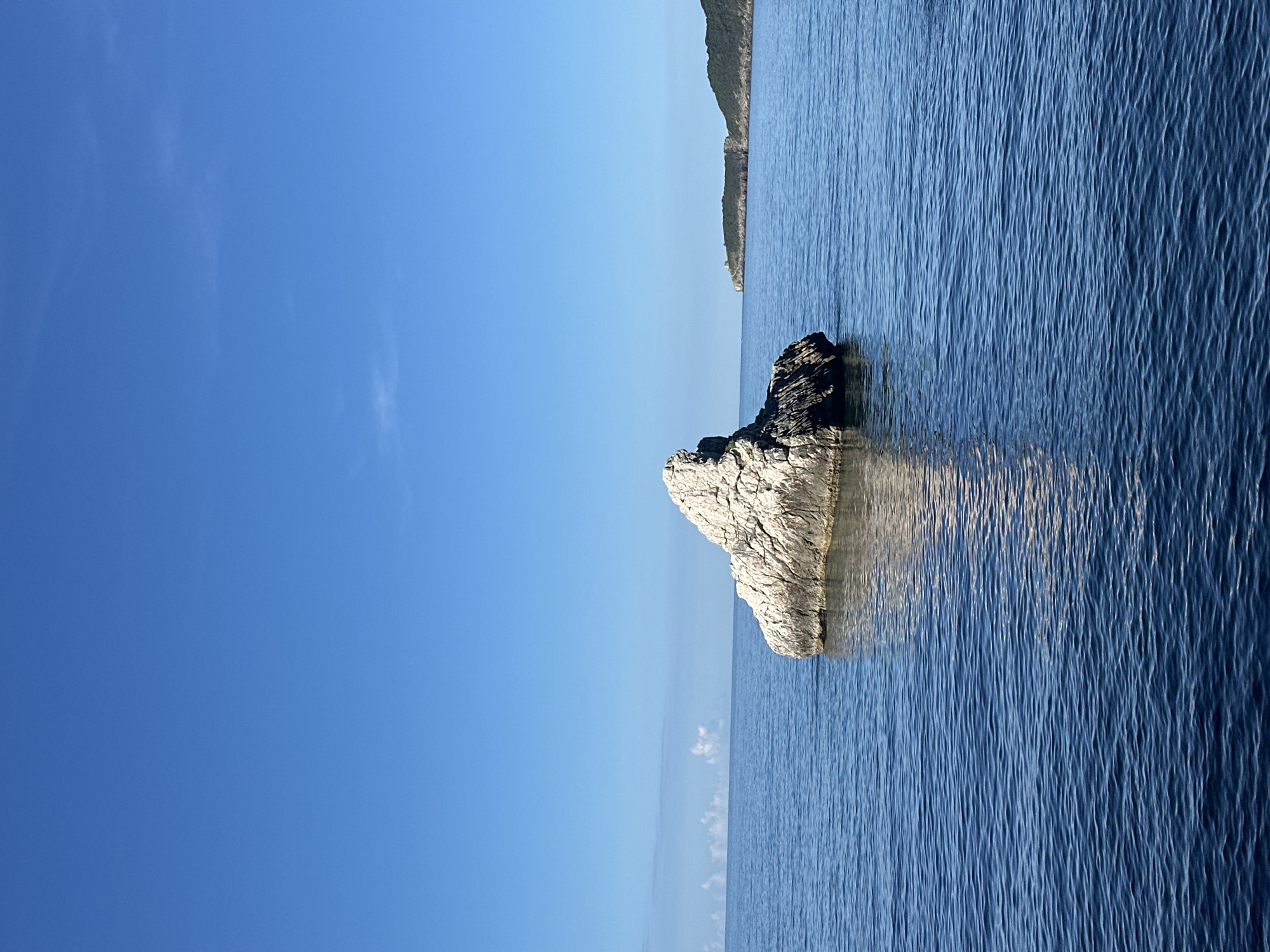 The image depicts a vertical view of a serene seascape. A large, jagged rock formation dominates the center of the frame, partially submerged in the tranquil blue water. The rock exhibits a mix of light gray and dark gray shades, suggesting varying exposure to elements and possibly different mineral compositions. The surface of the water ripples with subtle waves, creating gentle reflections around the rock. In the background, a distant landmass is visible, characterized by a hilly terrain covered in vegetation. The sky occupies a significant portion of the image, displaying a gradient of clear, cloudless blue. The overall atmosphere is calm and bright, indicating a sunny day. This scene depicts a location off the coast of Bečići, Montenegro.
