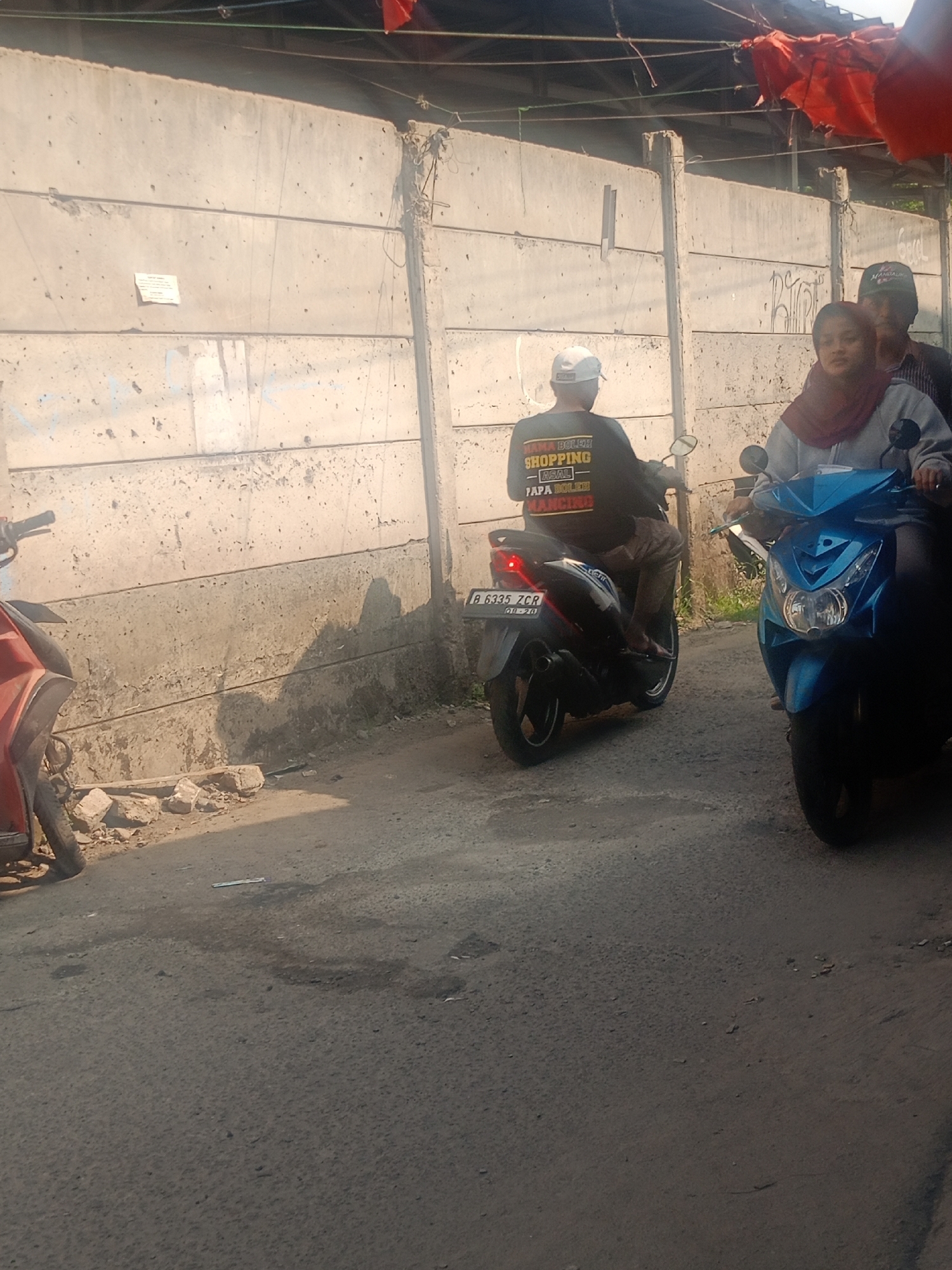 The image shows a street scene in Bojong Gede, Indonesia, with a concrete wall in the background and two motorcycles in the foreground. On the left, a red motorcycle is partially visible. In the center, a person wearing a black shirt with yellow text and a white cap is riding a black motorcycle. The text on the shirt reads "NAMA BOLEH SHOPPING - ASAL PAPA BOLEH NANCING". On the right, a blue motorcycle is ridden by a woman wearing a hijab and a grey jacket, with a man behind her wearing a green cap. The motorcycles are on a paved road. The environment appears to be an urban or semi-urban setting with a sunny day and shadows cast on the wall.