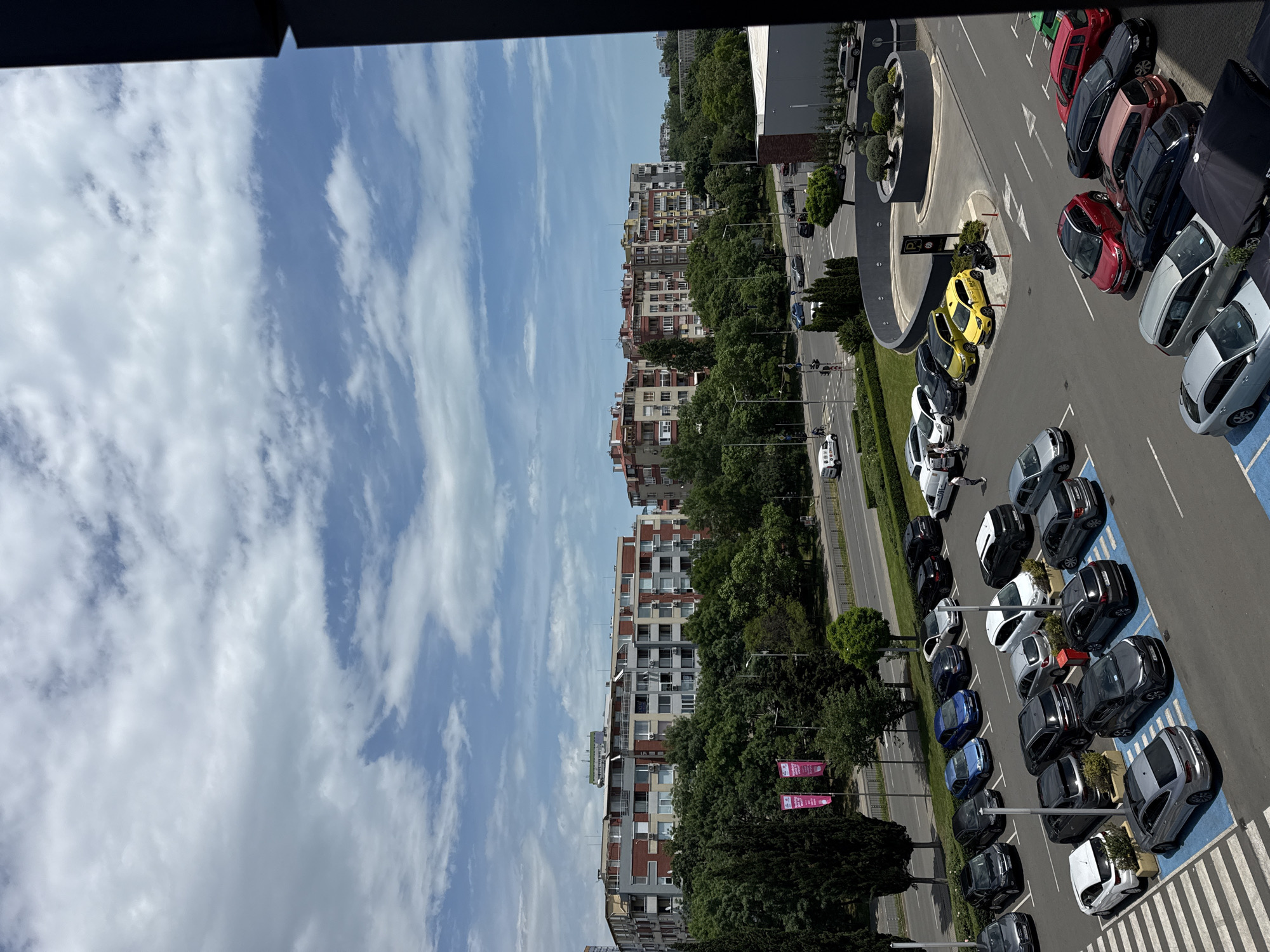 The image shows an aerial view of an urban landscape in Burgas, Bulgaria. In the foreground, there is a parking lot filled with various colored cars, including yellow, red, white, blue, and gray/black vehicles. The parking spaces are demarcated by blue paint, and small trees/bushes are planted at the end of some spaces.
Adjacent to the parking lot, a road runs parallel, lined with trees and buildings. The buildings are primarily residential apartments with red-tiled roofs and multiple stories. Beyond the buildings, there is a cloudy blue sky.
Traffic moves along the road. People are not clearly visible, but the presence of parked and moving cars suggests human activity in the area. The overall environment is urban with a mix of residential, commercial (parking), and green spaces.