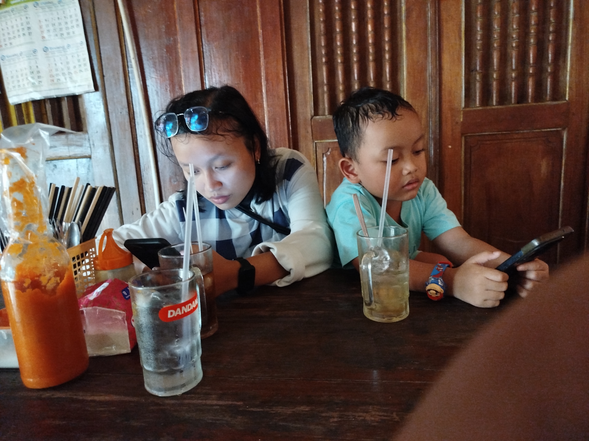 In the city of Pojok, Indonesia, two children are seated at a wooden table, engrossed in their mobile phones. The girl on the left, wearing glasses perched on her forehead, is looking down at her device. Beside her, a younger boy in a turquoise t-shirt is also focused on his phone. On the table are two glasses with straws, one appearing to contain a light-colored beverage, and another, partially obscured, holding a darker liquid. A bottle of red sauce or condiment stands to the left, and a basket of utensils is visible in the background. The scene is set against a backdrop of wooden paneled walls, with a calendar hanging on the left.
