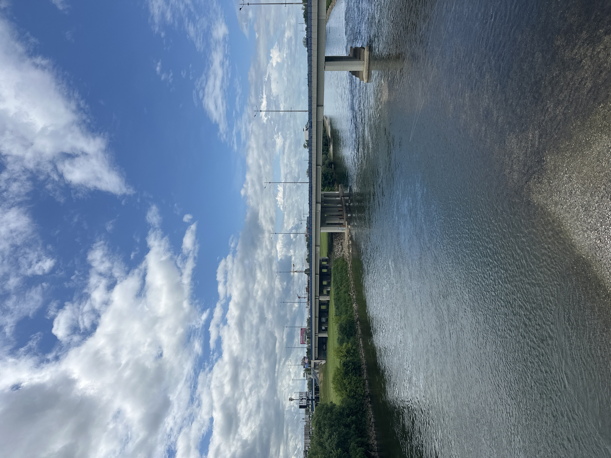 The image depicts a river scene in Zagreb, Croatia, with a bridge spanning across the water. The sky is filled with white, puffy clouds against a blue background. The river's surface reflects the sky, showing subtle ripples and a muted gray tone. The bridge is supported by concrete pillars. Trees and vegetation line the riverbank. The image is taken from a high angle, looking down onto the scene. The image is rotated 90 degrees clockwise.