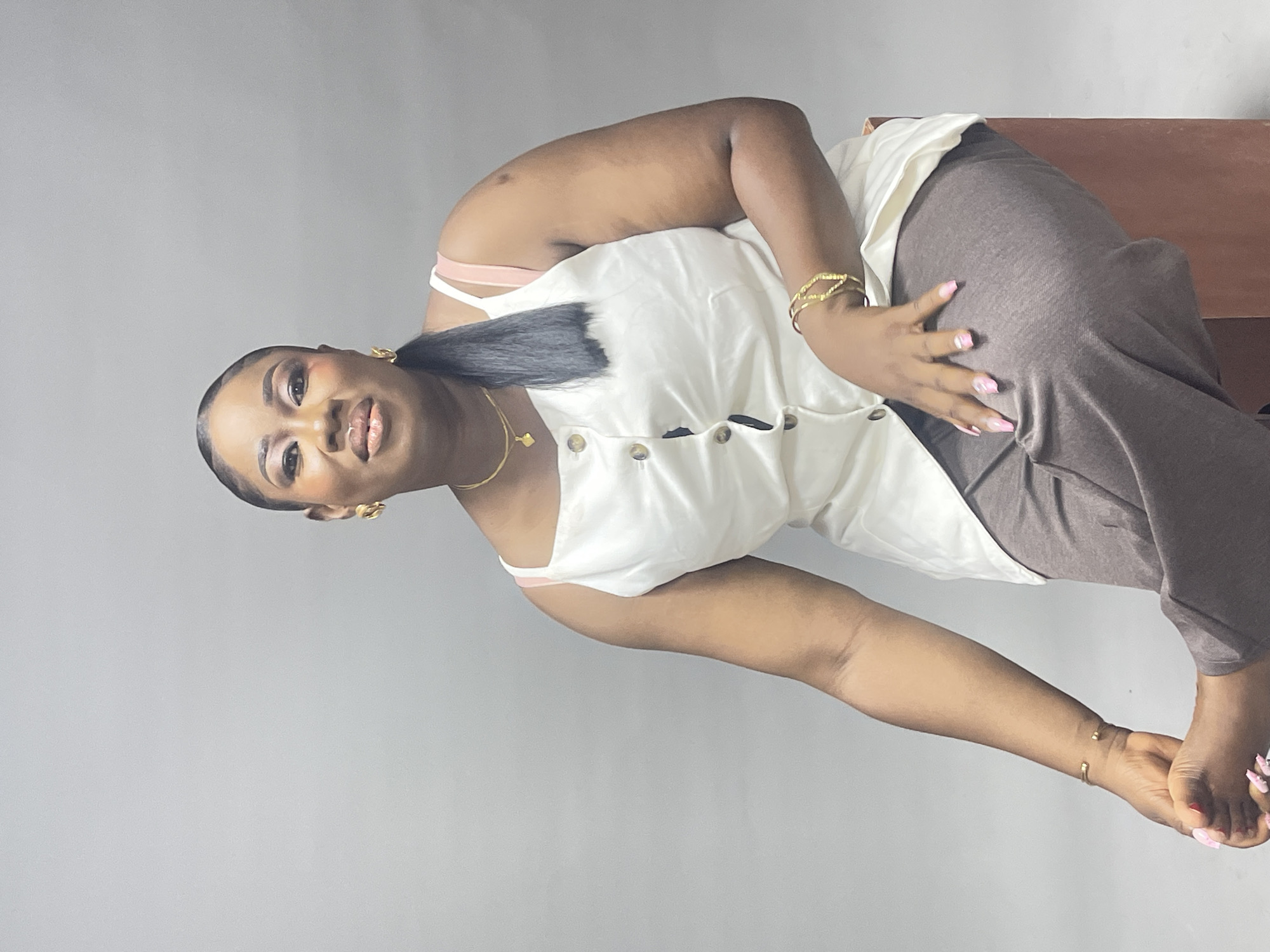 A woman is depicted indoors, likely against a neutral background. She is wearing a light-colored sleeveless top with button-down detailing, paired with brown trousers. The top appears to have thin pink straps. She is adorned with gold jewelry including earrings, a necklace, and bracelets on both wrists. Her hair is styled in a low ponytail. The woman's hand with manicured nails is placed on her hip. Location in Abuja, Nigeria, cannot be identified based on the image.