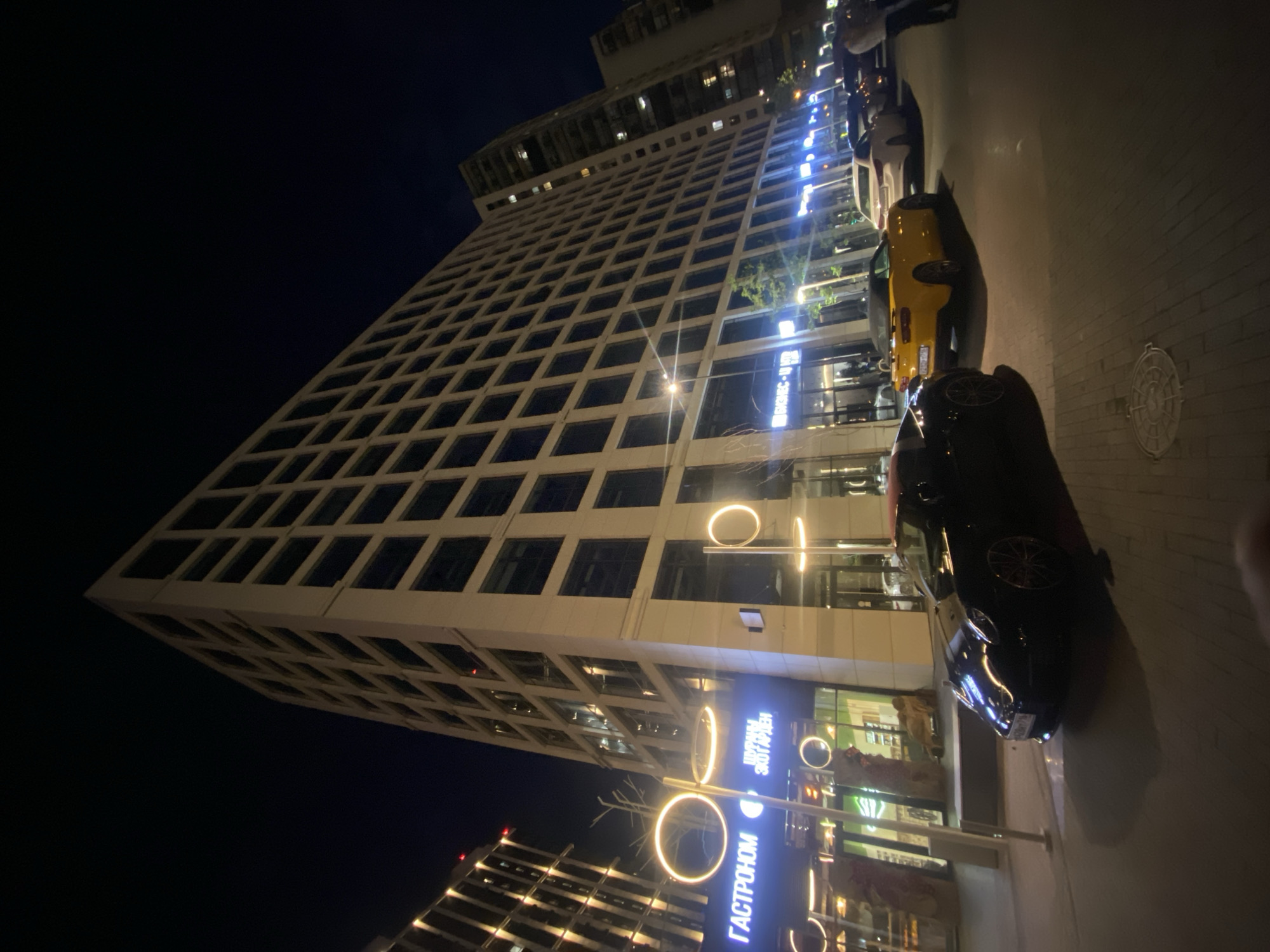The image depicts a nighttime urban scene in Kazan, Russia, showcasing modern architecture and street activity. A large, multi-story, light-colored building with a grid-patterned facade of rectangular windows dominates the left and central parts of the frame. Its ground level features several illuminated storefronts and two prominent signs displaying Cyrillic text: "ГАСТРОНОМ" (Gastronom) on the left and "ЭКОГАРДЕН" (Ekogarden) to its right.
Along the paved sidewalk in front of this building, several streetlights with distinct circular, glowing LED rings illuminate the area. Two luxury convertible sports cars are parked on the street, both facing towards the right: a black vehicle with a red soft top is in the foreground, followed by a bright yellow vehicle, both bearing Russian license plates. Further down the street on the right, additional parked cars, including a white sedan, are visible.
To the far left, a portion of another modern building with vertical light elements is visible. In the background on the far right, a tall residential-style building with multiple illuminated windows and balconies can be seen. One individual, wearing a light-colored upper garment, is observed walking away from the viewer on the far right edge of the frame. The street and sidewalk appear well-maintained.