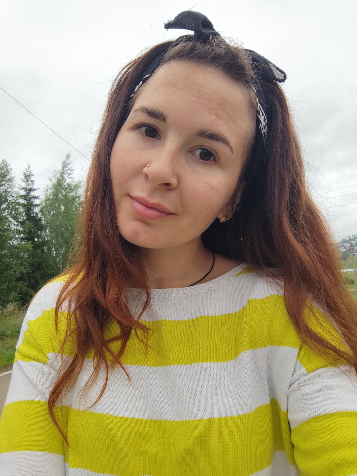 A young woman with reddish-brown hair, wearing a black bandana on her head and a white and yellow striped sweater, is captured in a close-up selfie. She has a small nose ring and a delicate earring in her left ear. The background suggests an outdoor setting, with blurred green trees and a cloudy sky, potentially indicating the outskirts of the city of Фоки, Russia, given the presence of distant houses and power lines visible.