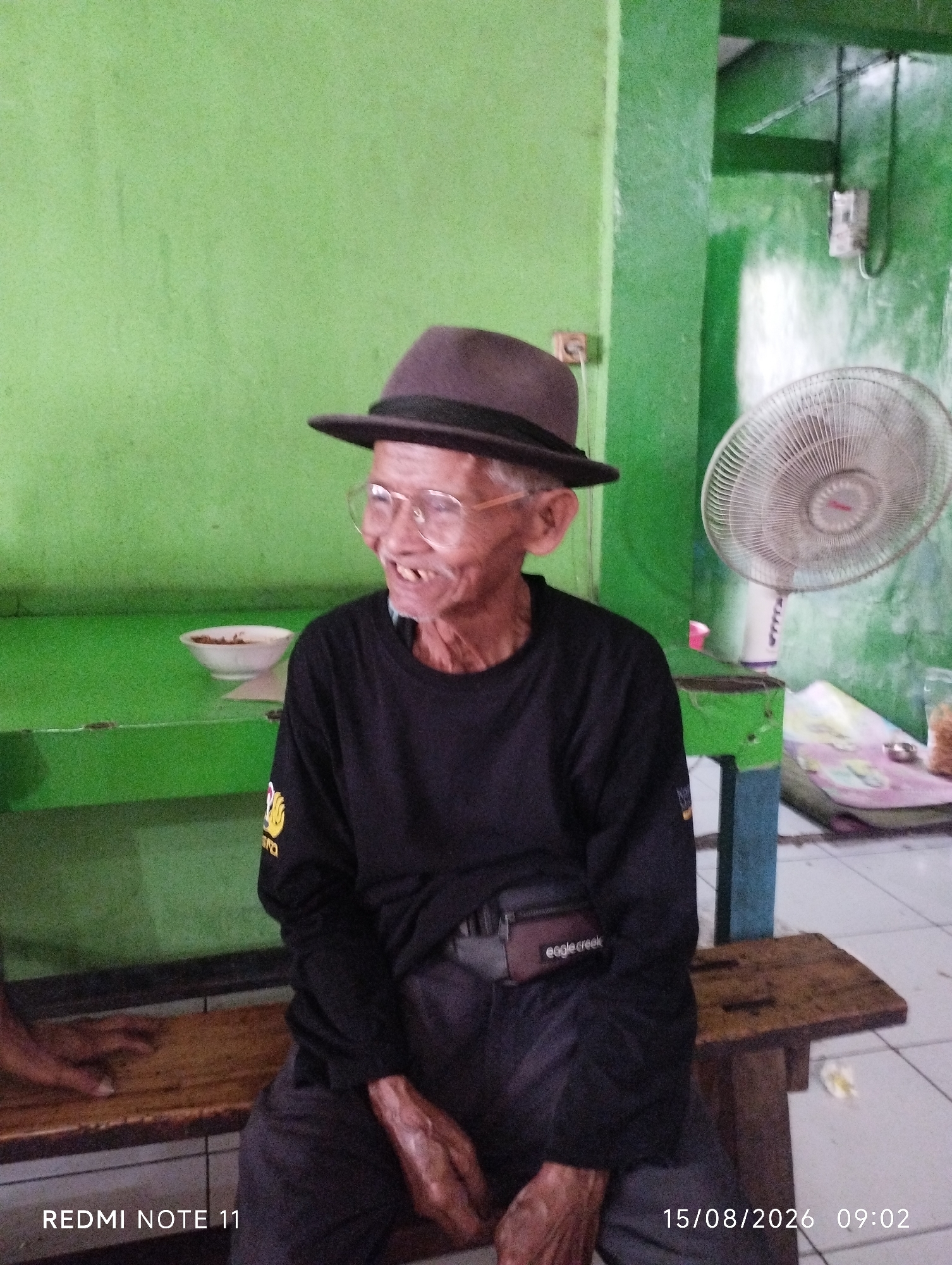 An elderly Indonesian man with glasses and a brown hat is seated on a wooden bench. He is wearing a black long-sleeved shirt and dark pants. A small pouch with "eagle creek" written on it is attached to his waist. The background is a bright green wall. A white bowl is on a table to the man's left, and a fan is visible in the background. The image is a photograph taken in Karawang, Indonesia.