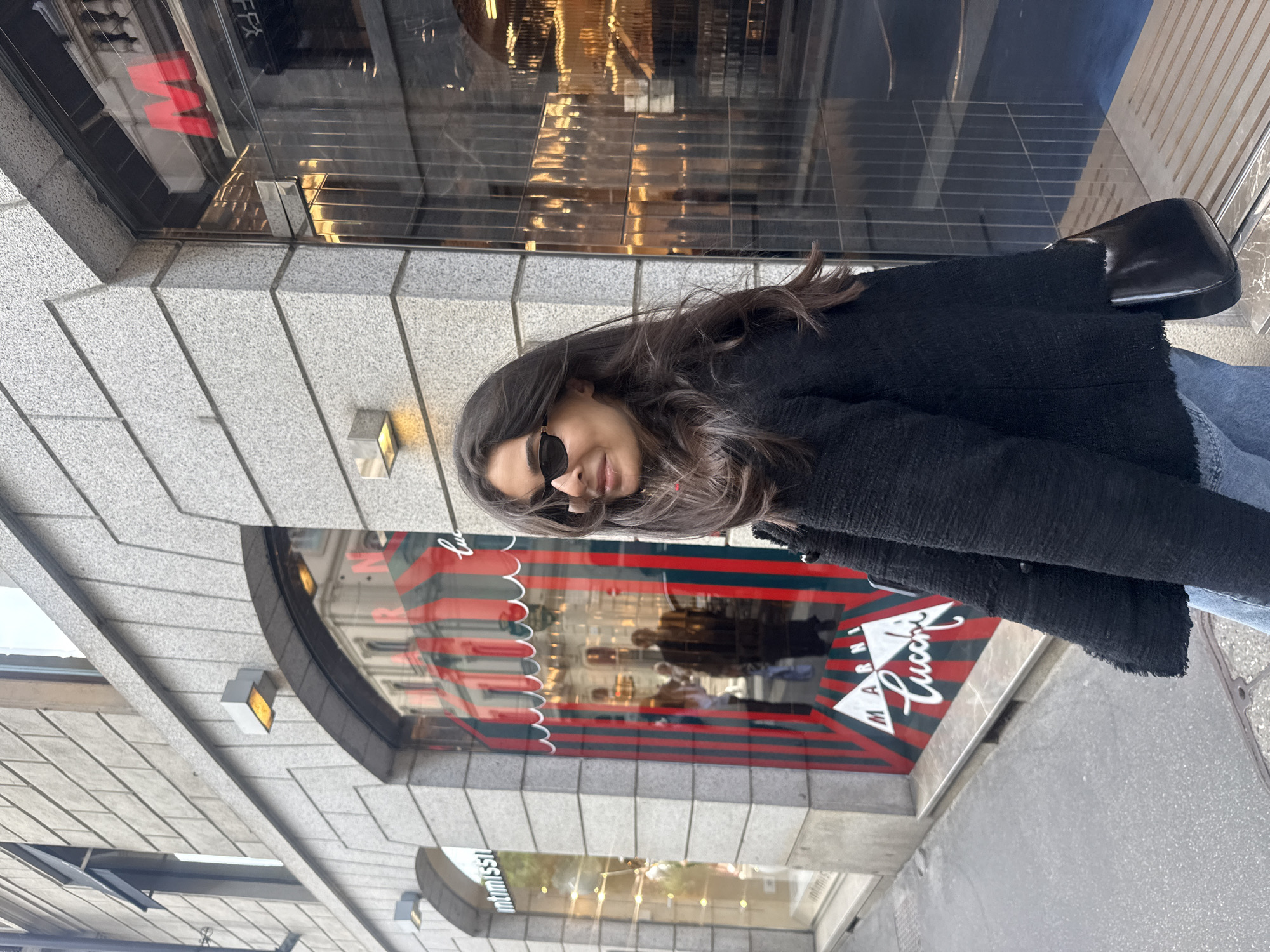 A woman wearing sunglasses and a black jacket is standing outside the Marni Lucci store in Milan, Italy. The store's window displays its logo. The building is made of grey stone, with storefronts visible above and below. The woman's shoulder bag is black.