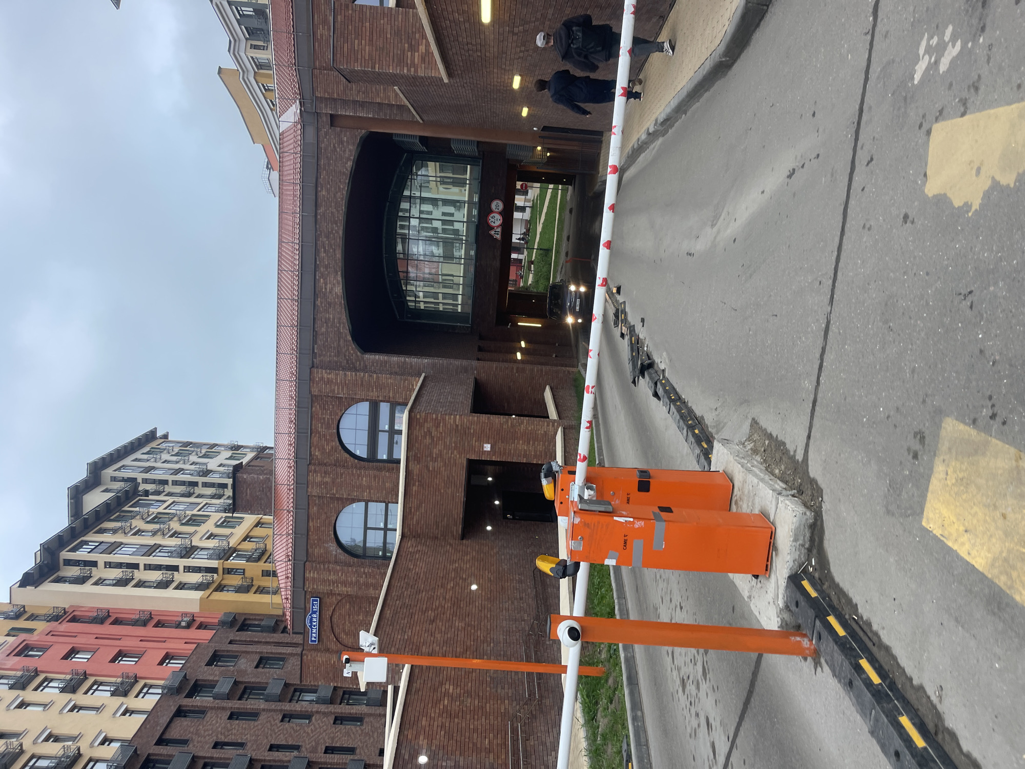 A ground-level, slightly tilted, outdoor shot depicts a barrier at the entrance to an arched brick structure, likely a parking garage, in Razvilka, Russia. The building on the left has multiple sections in different colors, including red, yellow, and brown. The address sign "РИМСКИЙ 15к1" (Rimskiy 15k1) is visible on the side of this building. The barrier has a white and red striped arm. Two orange posts hold up the barrier's mechanism, with yellow warning lights on top. A black vehicle with its headlights on is driving toward the camera in the distance, passing through the archway. Pedestrians are seen walking on the walkway behind the vehicle. The sky is overcast. The pavement in front of the barrier has yellow markings.