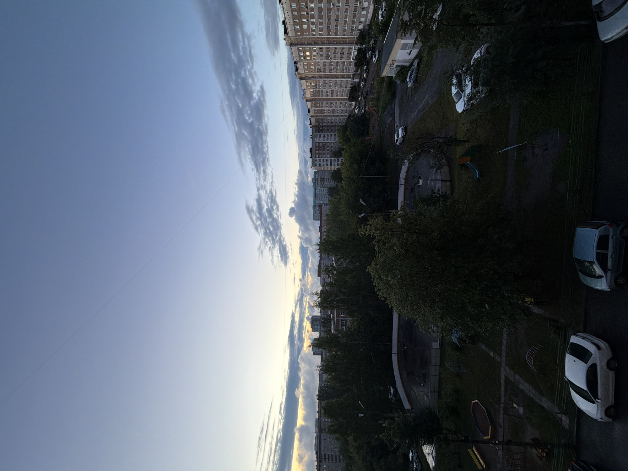 The image shows an aerial view of a residential area in Izhevsk, Russia, at what appears to be dawn or dusk. The sky is a gradient of light blue transitioning to yellow and white near the horizon, with scattered clouds. A thin, dark line, likely a power line or wire, stretches diagonally across the sky.
In the midground, a dense line of trees separates the foreground from a row of large, multi-story apartment buildings. The buildings are light-colored with many windows. Several cars are parked in front of and between the buildings.
The foreground features a courtyard or playground area with a dark circular surface, possibly a sports court or an ice rink. There are also playground structures visible, including a slide and a sandbox. Green grass and trees are scattered throughout the foreground. Several cars are parked in the foreground as well, including a white sedan and a silver hatchback. The ground in the foreground appears wet in places, suggesting recent rain. The scene depicts a typical urban residential environment.