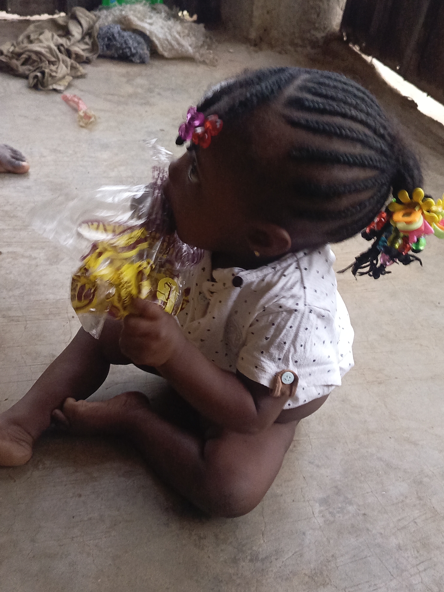 A young child with dark skin and braided hair sits on a concrete floor in Akwanga, Nigeria. The child wears a short-sleeved white shirt with dark polka dots and colorful hair decorations. The child is holding a plastic bag containing yellow and purple objects. The background includes miscellaneous items and a partially visible wall.