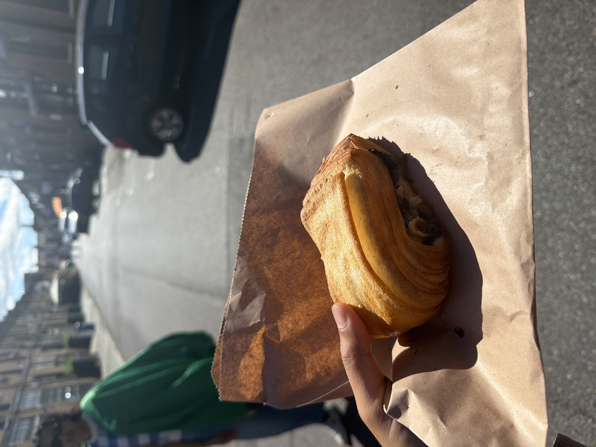 A person's hand holds a brown paper bag containing a pastry, possibly a pain au chocolat, in the foreground. The pastry is golden brown and flaky, with visible layers and a dark filling. The background shows a street scene in what appears to be London, United Kingdom, with buildings lining both sides of the road. A black car is parked on the right side of the street. A person wearing a green garment is visible on the left side of the image. The sun is shining, casting shadows on the street.