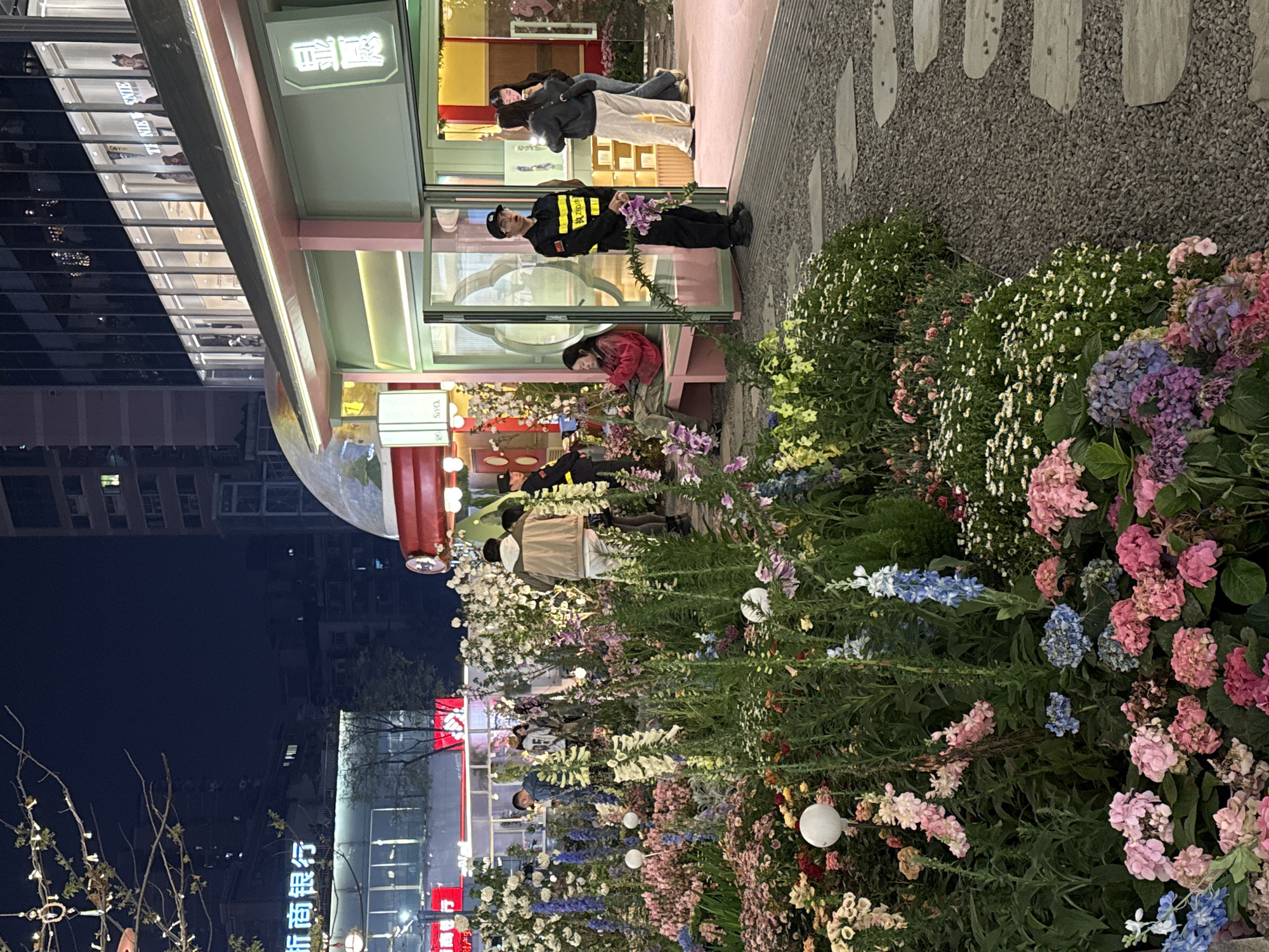 The image depicts an illuminated outdoor commercial area at night in Liangjiang, China, dominated by an extensive decorative flower garden. In the foreground and mid-ground, a dense arrangement of various flowering plants, including hydrangeas, delphiniums, and daisies, in shades of pink, blue, purple, white, and yellow, occupies the center. Multiple spherical white lights are interspersed within the flower beds. To the immediate right, a gravel path with irregular grey stepping stones leads towards a light mint green building with pink accents and large glass doors.
Seven individuals are clearly visible. A male security guard in a black uniform with reflective yellow stripes and a black cap stands at attention to the right of the building's entrance, holding a small bouquet of purple flowers. Seated on a light pink bench inside the building's open entrance, a female in a red jacket and olive green pants is looking at her mobile phone. Two additional females stand within the entrance: one in a dark top and light pants is looking at her phone, while the other in a dark top and jeans faces her. Further back, amidst the flowers, a male in a light brown jacket and white pants walks away from the viewer. Other indistinct figures are present further into the scene.
The building on the right features a glowing sign with Chinese characters ("鼎盛") above its entrance. Above this low-rise structure, a multi-story building with dark glass reflects internal lighting, with a partially visible "TINIE WINIE" sign on its facade. In the background to the left, a large commercial building displays "浙商银行" (Zheshang Bank) in illuminated Chinese characters, with other brightly lit signs and building structures visible in the distance. Tree branches adorned with string lights are in the upper left portion of the image. The scene is artificially lit, suggesting an evening setting.