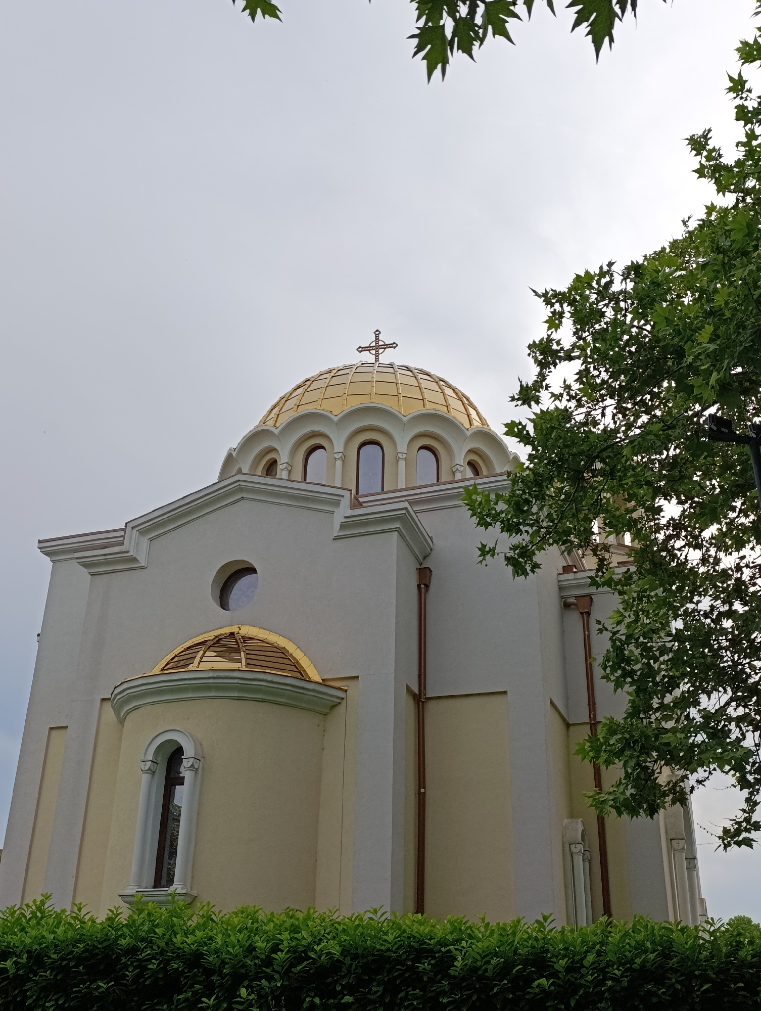 A low-angle, medium shot captures the facade of a cream-colored, modern-style church with a prominent golden dome topped with a cross. The dome features arched windows and is surrounded by white decorative molding. A smaller golden semi-dome is visible on the lower left, also with an arched window. Green foliage frames the bottom of the image, and a lush green tree with broad leaves partially obscures the right side of the church and the sky. The sky is overcast and pale gray. There are no people or animals present in the image. The scene is static and depicts the exterior architecture of a religious building.