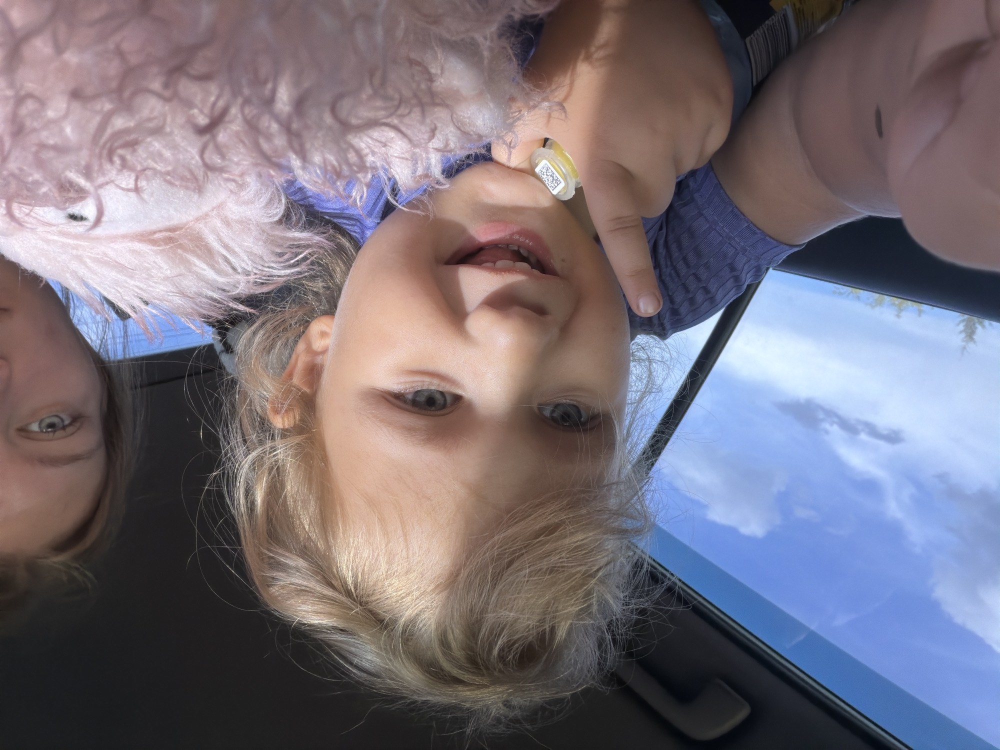 A close-up selfie shows a young child with blond hair and blue eyes in the foreground, holding a stuffed animal. The child is wearing a purple shirt and has a yellow ring on their finger with a QR code. Behind the child, a young woman with blond hair is visible, looking towards the camera. The scene appears to be inside a car, with a blue sky and clouds visible through the window. The location is identified as Novoe Mavrino, Russia.