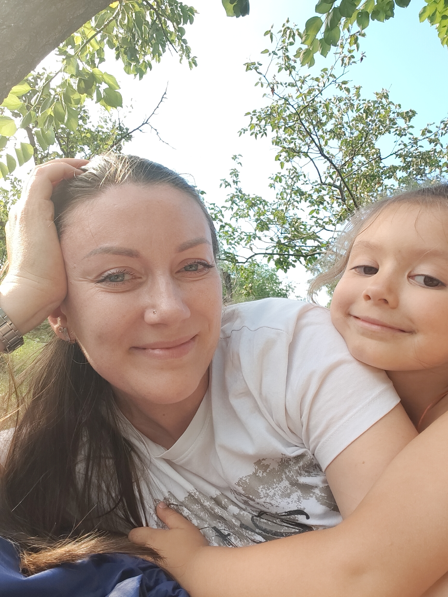 The media file is a selfie taken outdoors, likely in a park or garden given the presence of trees and foliage. Two people are visible: an adult woman and a young child. The woman is in the foreground, her left hand resting on her head. She has long, dark brown hair, light skin, and blue-green eyes. She is wearing a light-colored t-shirt with a graphic design. The child is nestled close to the woman, with their head resting on the woman's shoulder. The child has light brown hair, light skin, and brown eyes. They are also wearing a light-colored t-shirt. Both subjects appear to be smiling and in a relaxed, affectionate pose, suggesting a moment of connection and enjoyment.
The setting appears to be daytime, with bright sunlight filtering through the leaves of the trees, creating dappled lighting. The sky is a clear blue, indicating fair weather. The overall mood is warm and joyful. The image is a close-up shot, focusing on the faces and upper bodies of the two individuals. There are no visible objects that would provide strong clues about the specific location within Kryvyi Rih, Ukraine, beyond it being an outdoor, natural setting. The image appears to have been taken in the summer or late spring due to the lush green leaves on the trees.