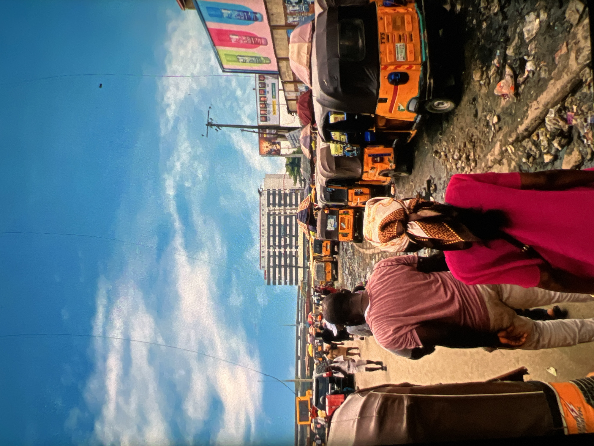 The image depicts a busy street scene in Lagos, Nigeria, likely a market area. The sky is a bright blue with scattered white clouds. A row of orange auto rickshaws, also known as Keke Napep, are parked along the right side of the street. The road surface is rough and uneven, with debris scattered on the ground. In the background, several multi-story buildings are visible, including one with a distinct grid-like facade. Other vehicles and market stalls with colorful awnings are also present, indicating commercial activity. People are walking along the street, some appearing to be customers and vendors. The overall impression is one of a vibrant and bustling urban environment.