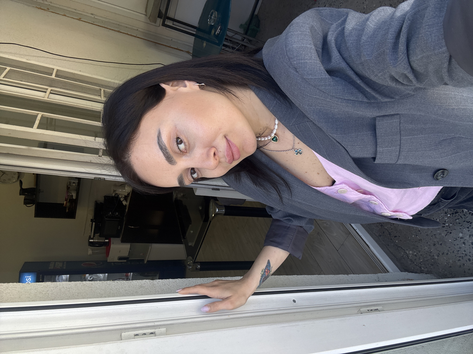 A person with dark hair is taking a selfie by a window or doorway. The person is wearing a gray blazer over a pink collared shirt, and a pearl necklace. A butterfly tattoo is visible on the forearm. The person is touching the white frame of the window. Behind the person through the window/door, there appears to be a business interior with a Pepsi-branded cooler. The location is likely Tbilisi, Georgia, based on the person's appearance. The scene seems casual.