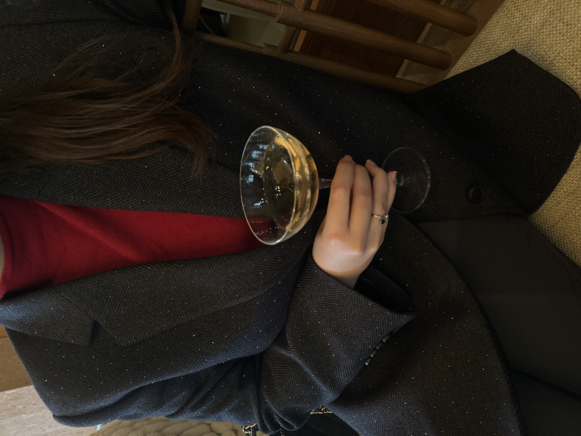 A person is holding a coupe glass filled with a light-colored liquid. The person is wearing a red garment beneath a dark herringbone-patterned coat with subtle glitter-like accents. A ring is visible on the person's finger. The environment appears to be an indoor setting. The location is unknown and cannot be determined to be in Yerevan, Armenia, based on the image alone.