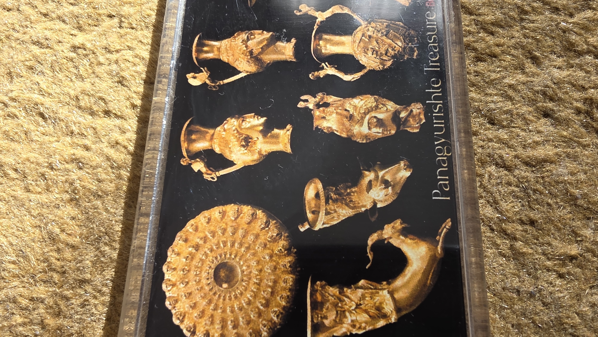 The image shows a framed photograph or a printed image of the Panagyurishte Treasure, a collection of Thracian golden artifacts. The artifacts, depicted in a gold hue against a black background, include several rhyta (drinking horns) in the shape of animal heads and a phiale (a shallow dish). The text "Panagyurishte Treasure" is visible on the right side of the frame. The photograph or image is placed on a textured, brown surface. There is no information about the location in the city of Blagoevgrad, Bulgaria or other details about people, subjects, objects, actions, interactions, and the environment.