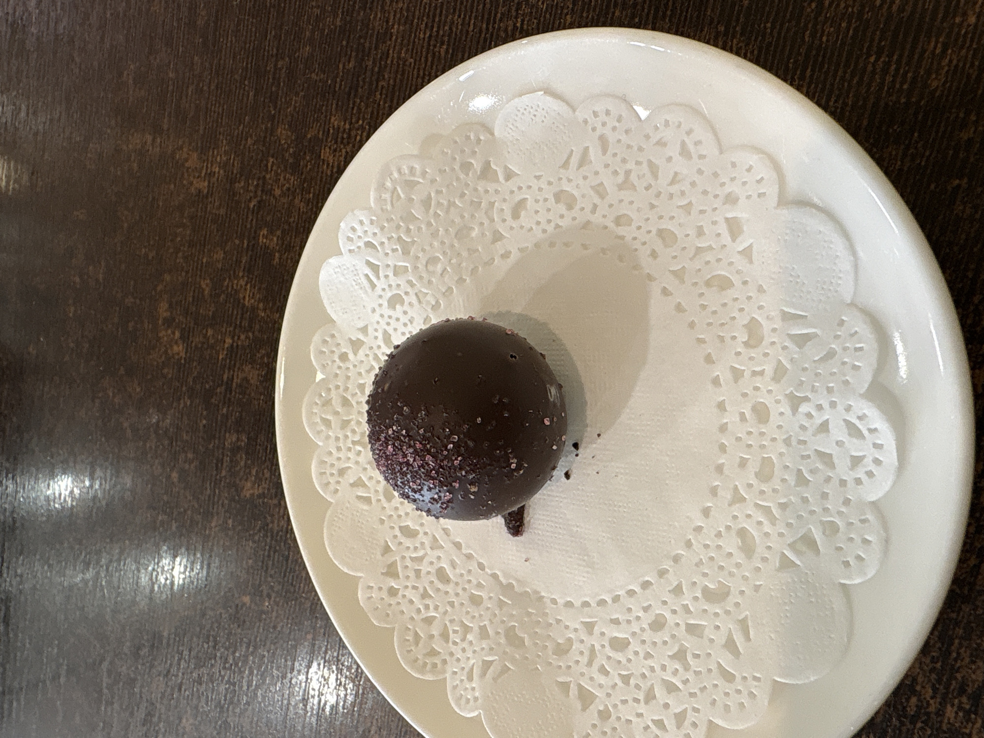 The image shows a single chocolate truffle sprinkled with pink sugar on a white paper doily, placed on a white plate. The plate rests on a dark wooden surface. The lighting suggests an indoor setting, possibly a cafe or restaurant. No specific location within Kaliningrad, Russia can be determined from the image. There are no people or actions depicted.