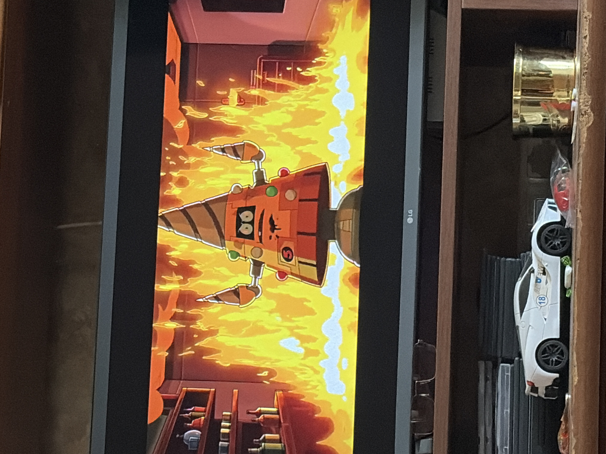 An animated robot with a conical head and orange body, decorated with green and red circular accents, stands engulfed in flames. The robot has drill-like appendages extending from its sides and a distressed facial expression with black eyes and a mouth. It is positioned in a room with wooden shelves holding bottles on the left and shelving units on the right. The background is dominated by intense orange and yellow flames, suggesting a fiery environment. Below the television screen, a white toy car with the number 18 and the logo "AIR-DEW" on its side is visible. Further to the right, a golden cylindrical container rests on a wooden shelf. The overall scene is captured from a slightly downward angle, suggesting the viewer is observing a television screen from a room below.