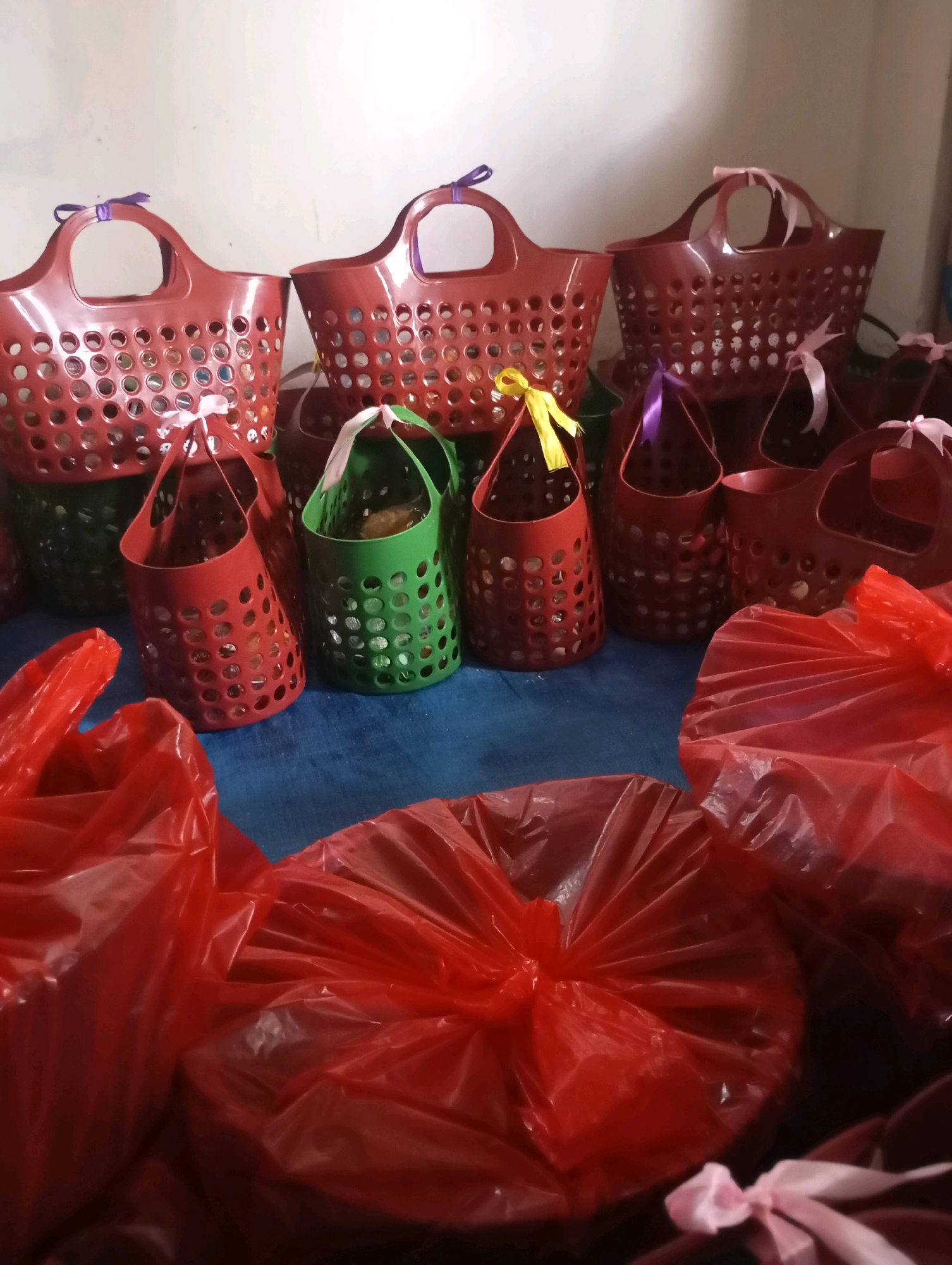 The image depicts a collection of red and green plastic baskets, some with purple, pink, or yellow ribbons tied to their handles. Several of these baskets appear to be filled with items. In the foreground, two large, crinkled red plastic bags, also tied with pink ribbons, are prominently displayed, obscuring the lower part of the baskets behind them. The scene is set against a light-colored wall in what appears to be an indoor setting. The overall arrangement suggests preparation for an event or distribution, possibly as gifts or favors. This scene is located in the city of Sumberjati, Indonesia.