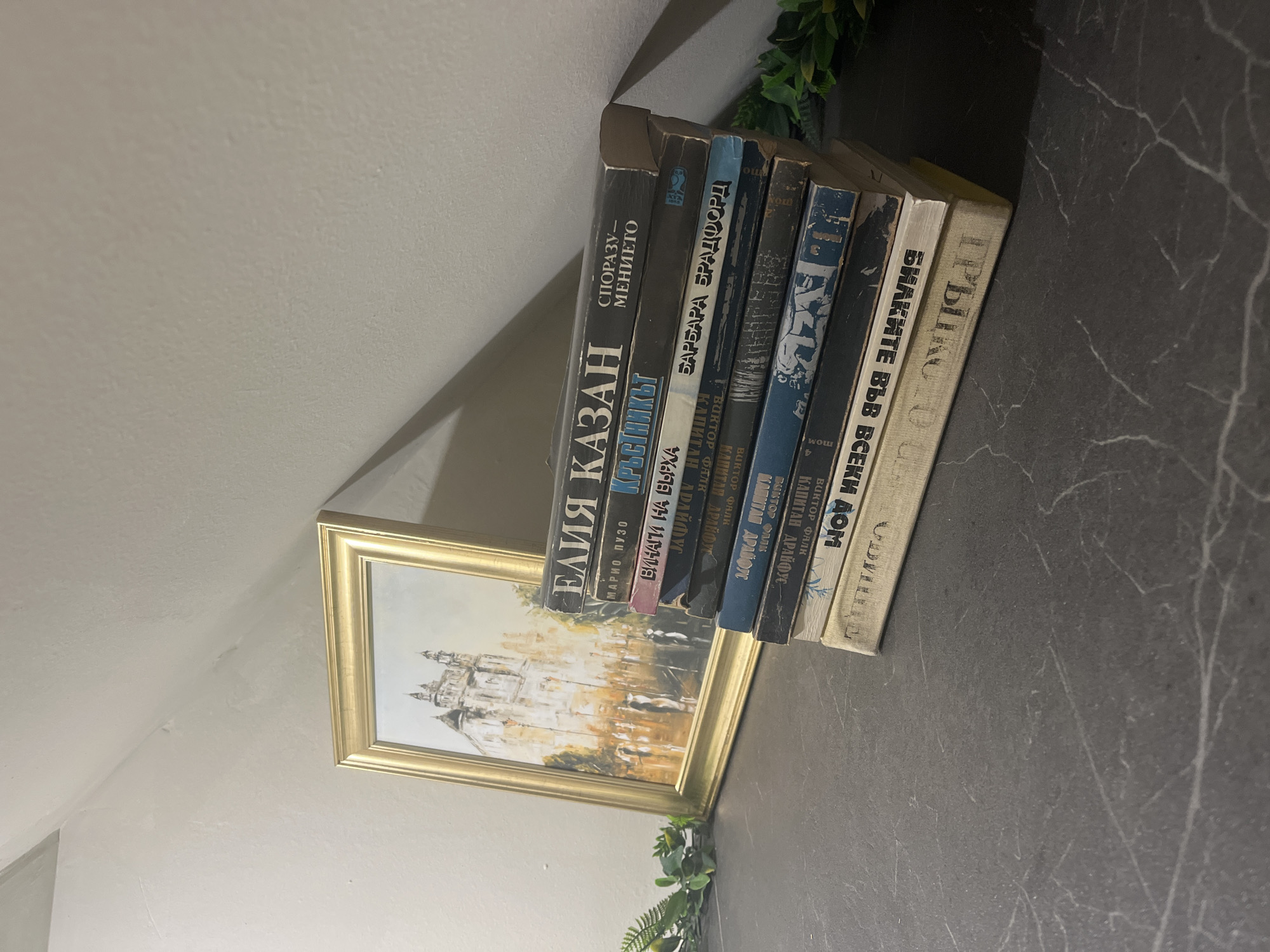 A medium shot shows a stack of six Bulgarian-language books leaning against a framed picture of a building on a dark gray countertop with white veins. The books are arranged vertically, their spines facing the viewer. From left to right, the titles visible are "ЕЛИЯ КАЗАН" by Mario Puzo, "КРЪСТНИКЪТ," "ВИНАГИ НА ВЪРХА," "БАРБАРА БРАДФОРД," "КАПИТАН ДРАЙФУС," and "БИЛКИТЕ ВЪВ ВСЕКИ ДОМ." The framed picture is a watercolor painting of a cathedral in a golden frame. Green foliage decorates the scene. The setting appears to be indoors, with a white wall and ceiling visible in the background. The location is unknown.