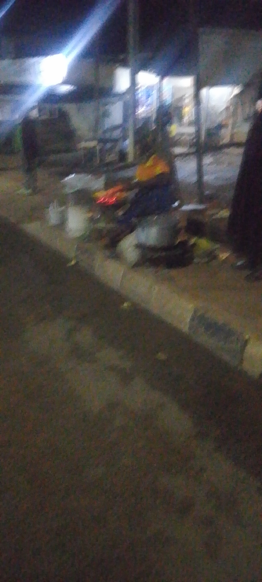 The image depicts a dimly lit, outdoor scene at night, likely in Gura Top, Nigeria. In the foreground, a textured, dark asphalt surface dominates, with a concrete curb running along the right side. The curb is partially obscured by shadows and appears to be wet in places, reflecting some light.
The middle ground reveals a more active area. Several individuals, their features indistinct due to the low light and motion blur, are present. One person is wearing a bright yellow garment and dark blue trousers, and appears to be interacting with objects that are not clearly identifiable but may include containers or food items. Another individual, wearing a dark outfit, stands further back. There are also various scattered objects, including what appear to be buckets or bins. Red lights, possibly from a vehicle or an appliance, are visible in the mid-ground.
The background shows the illuminated facade of a building or storefront. Bright lights emanate from within, casting reflections on the wet ground. The overall atmosphere is that of a street scene at night, possibly a market or a gathering area, characterized by low visibility and a sense of activity. The image suggests a public space where people are present and engaged in activities under artificial lighting.