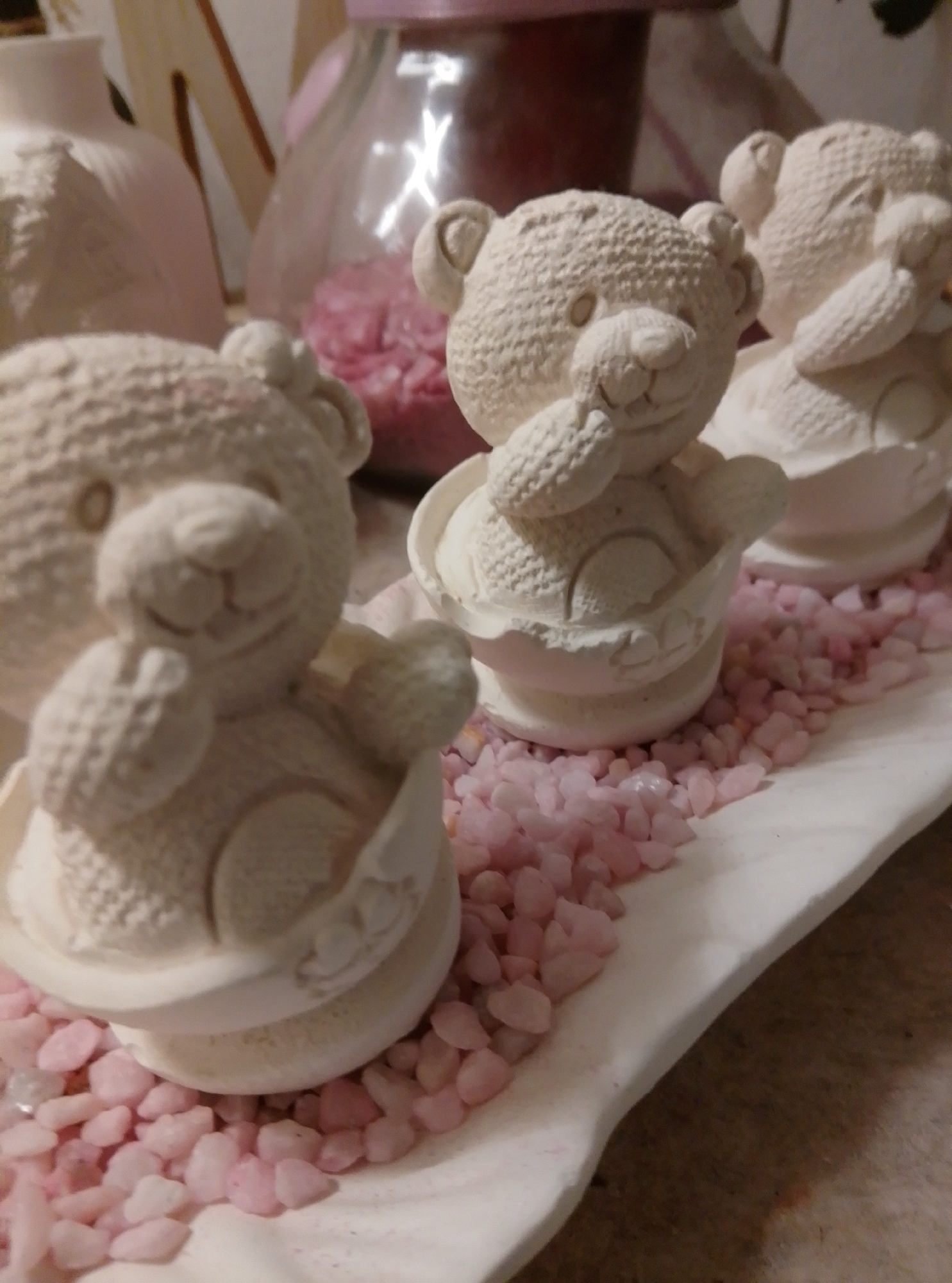 Three unpainted ceramic bear figurines are displayed on a white platter. The bears are arranged in a row, with the closest bear in the foreground on the left. Each bear is sitting in a small, rounded, bowl-like base and has its paws raised to its mouth. The platter is covered in small, light pink gravel. In the background, out of focus, are other decorative items including a large pinkish vase and a glass jar filled with more pink decorative material. The scene is likely indoors, possibly on a table or shelf. This image was taken in Wedel, Germany.