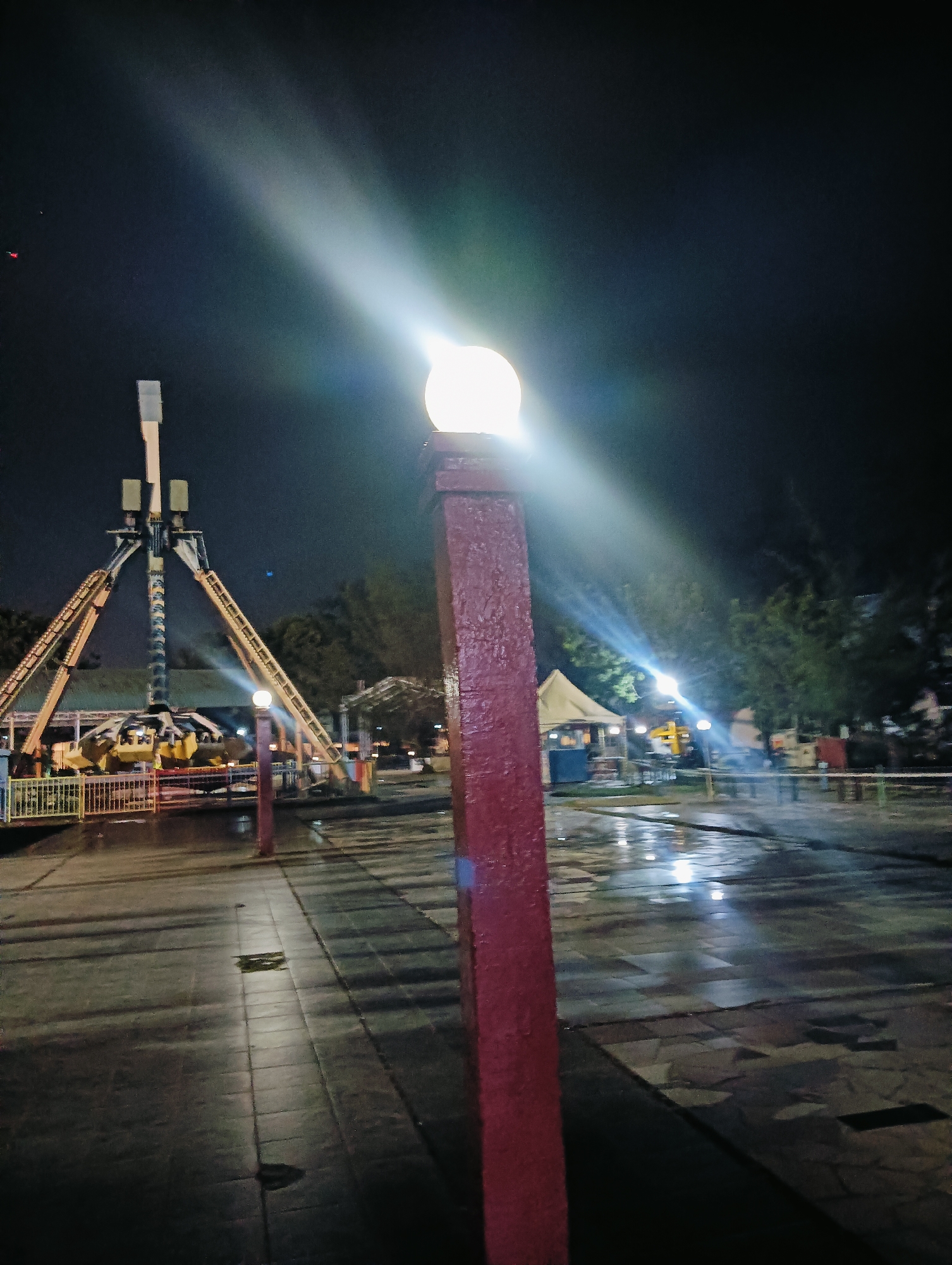 The image shows an amusement park at night in Batam, Indonesia. A tall pendulum ride, likely the "Bungee Trampoline," is visible in the background. The ground is wet, suggesting it recently rained, and reflects the lights from the park's attractions. In the foreground, a red pillar with a light fixture is prominently featured, casting a bright beam into the dark sky. The park appears to be closed or has few visitors, creating a quiet and atmospheric scene.