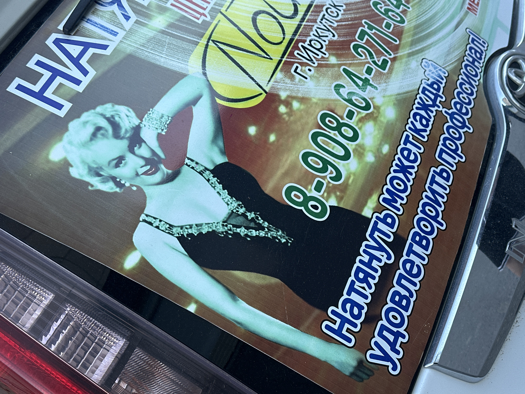 The image features a sign partially visible above a car's taillight. The sign displays a photograph of Marilyn Monroe, a phone number (8-908-64-271-64), and text in Russian. The text includes the name "Natalya" and the city "Irkutsk." The overall aesthetic appears dated and stylized, potentially advertising a service or business. The image's angle suggests it was taken in close proximity to the subject. The presence of the phone number, and the phrase "Natalya," along with the city Irkutsk, suggests that the location in Sayansk, Russia, may be related to the content of the sign.