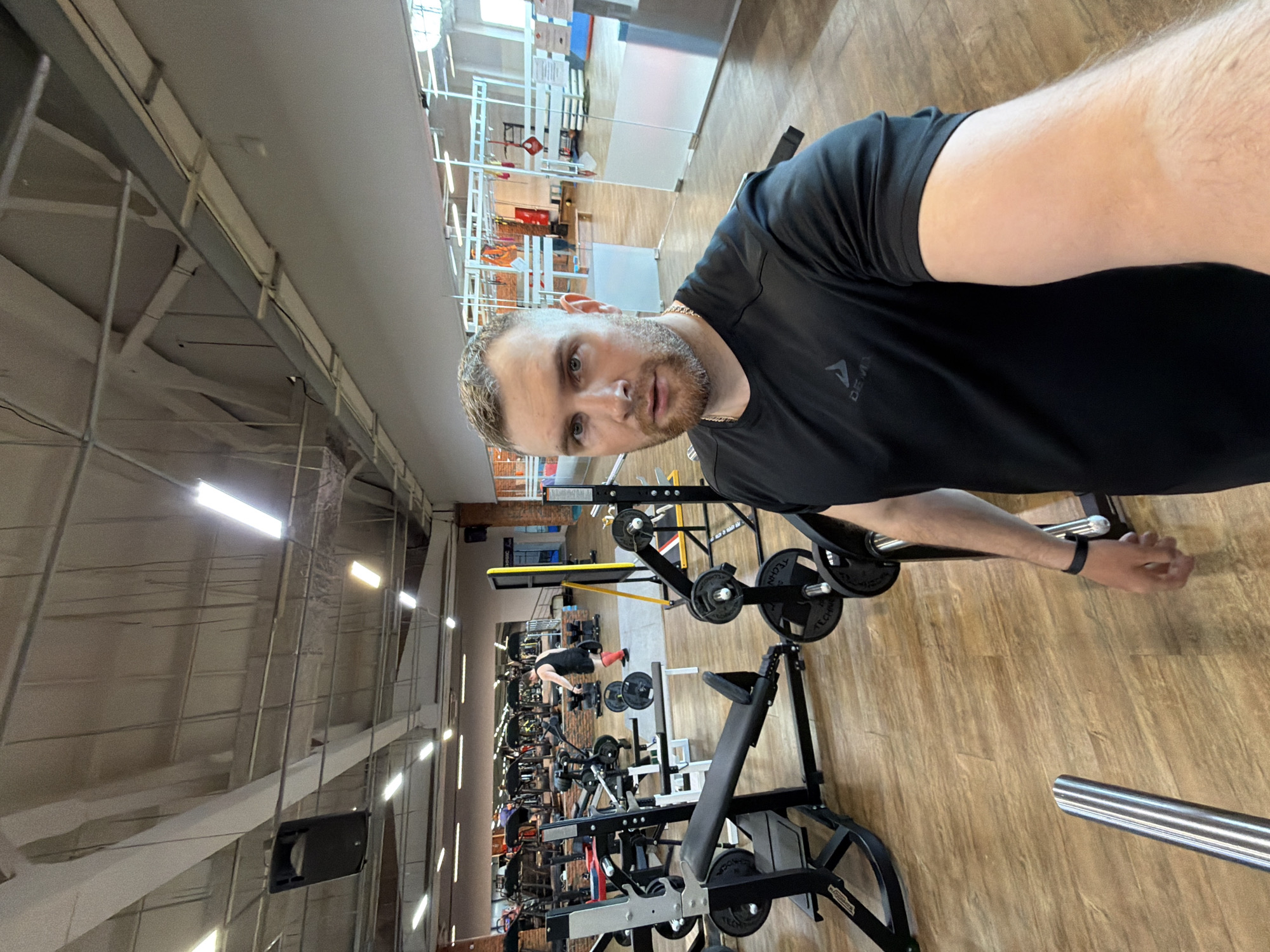 A man in a black t-shirt is posing in a gym. He has short blonde hair and a beard. He's standing near weightlifting equipment. Other people are exercising in the background. The gym has a high ceiling with exposed metal beams and fluorescent lighting. The floor is light wood. Based on metadata, the image was taken in Chaikovsky, Russia.