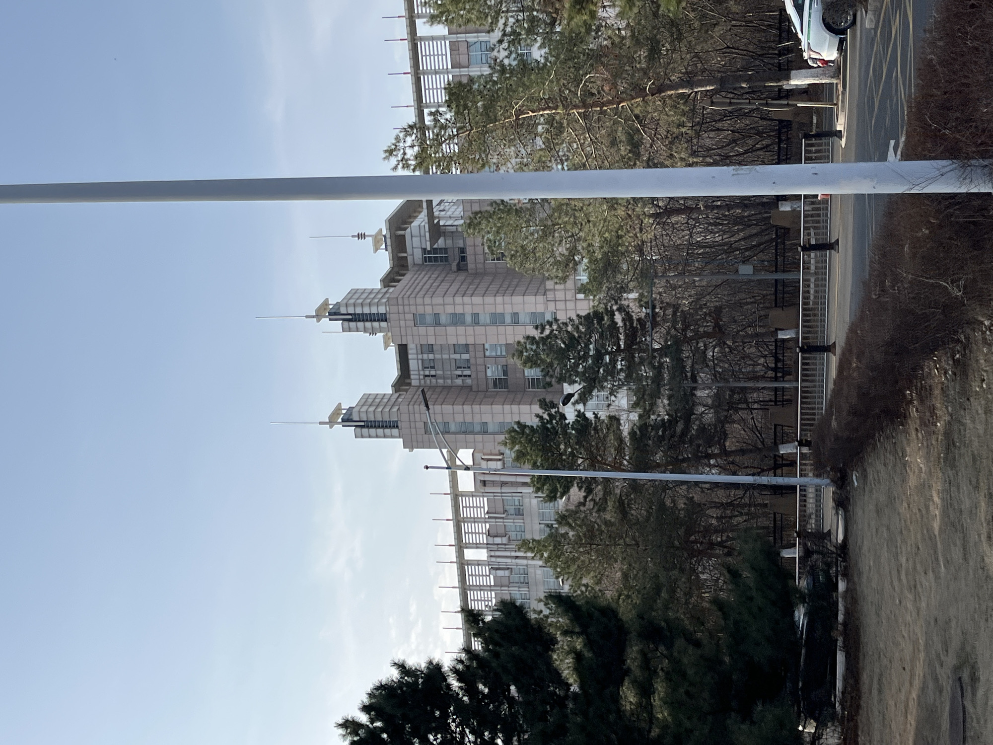 The image depicts a multistory building in Changchun City, China, partially obscured by trees and a utility pole. The building features a light-colored facade, multiple stacked sections, and antenna-like structures on its roof. There is a street with a yellow line and a portion of a car visible on the right side of the image. The foreground includes dark green pine trees and an area covered in brown foliage. The sky is clear blue.