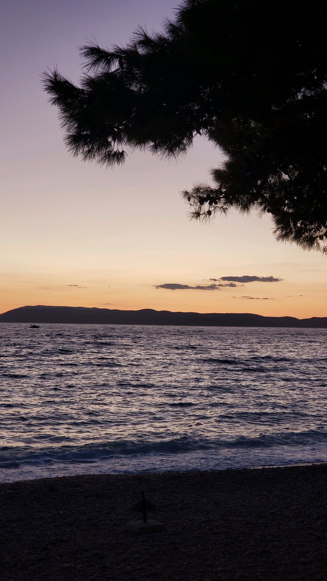 This media file depicts a serene coastal scene at dusk, likely in Općina Tučepi, Croatia. The sun has set, casting a soft gradient of purple and orange hues across the sky. Silhouetted against this tranquil backdrop is the dark, jagged outline of a pine tree branch with its needles prominently displayed, framing the top portion of the image.
Below the sky, the calm sea stretches out, its surface rippled by gentle waves. The water reflects the muted colors of the sky, appearing in shades of deep blue and purple. In the distance, the dark, mountainous silhouette of a coastline is visible, with a small boat subtly present on the water, adding a sense of scale and isolation to the vast expanse.
The foreground is a pebble beach, rendered in dark tones due to the low light. A small, dark object, possibly a part of a beach umbrella stand or a decorative element, is visible on the beach, anchored by a concrete base.
The overall mood is one of peace and quietude, characteristic of a seaside evening after the day's activities have concluded. The time of day is clearly sunset or twilight, with the fading light contributing to the subdued color palette and the sense of calm. There is no visible text in the image.