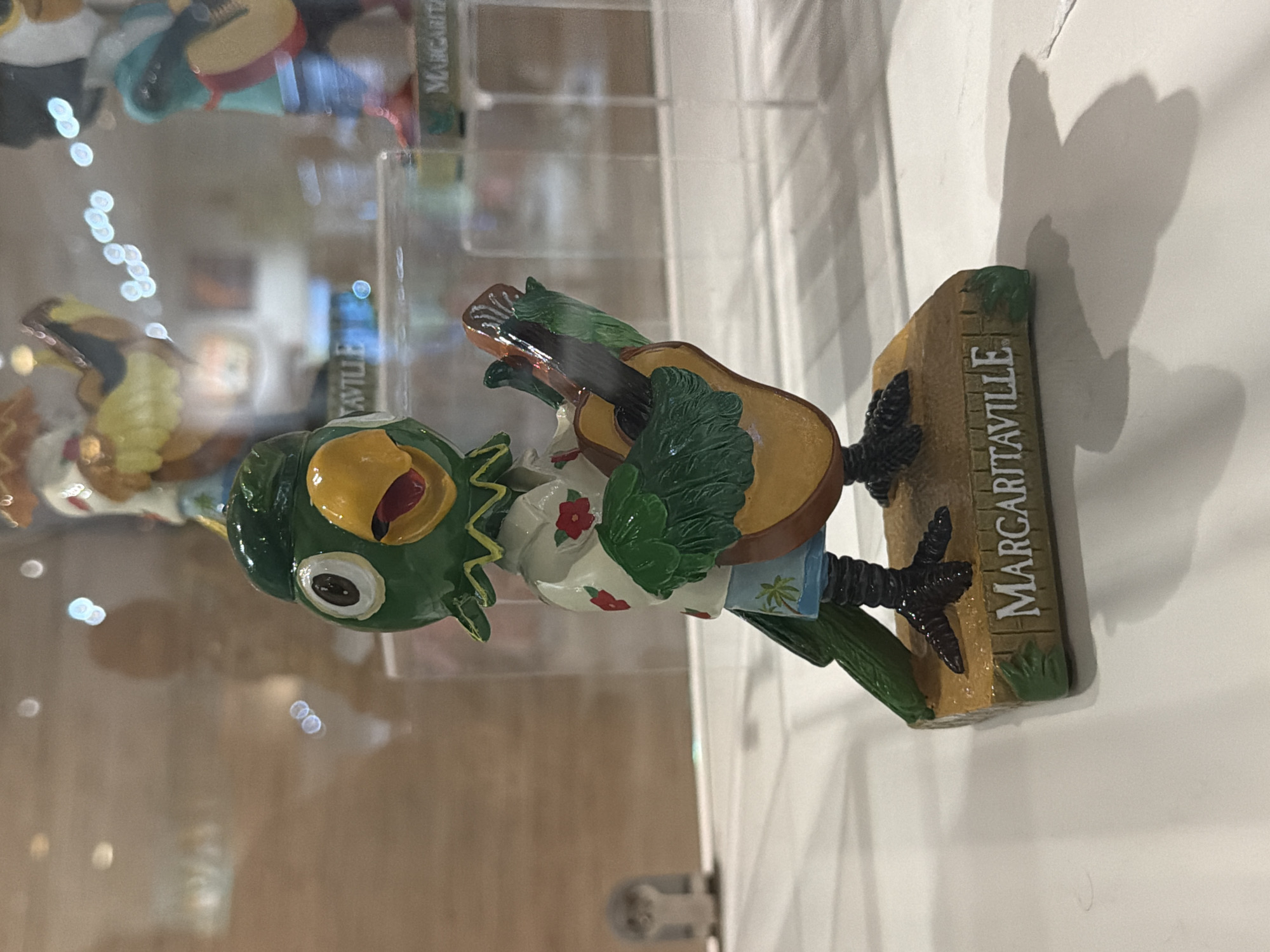 A close-up shot shows a decorative figurine of a green parrot playing a guitar. The parrot is wearing a white shirt with red floral patterns and blue shorts with a palm tree design. It stands on a wooden base with the word "MARGARITAVILLE" inscribed on it. The figurine is placed on a white surface, possibly a display shelf, with a blurred background that suggests an indoor environment, possibly a store or museum, with reflections of lights and other objects visible. The lighting is soft, casting a shadow of the figurine to the right. The image quality is slightly blurry, especially in the background.