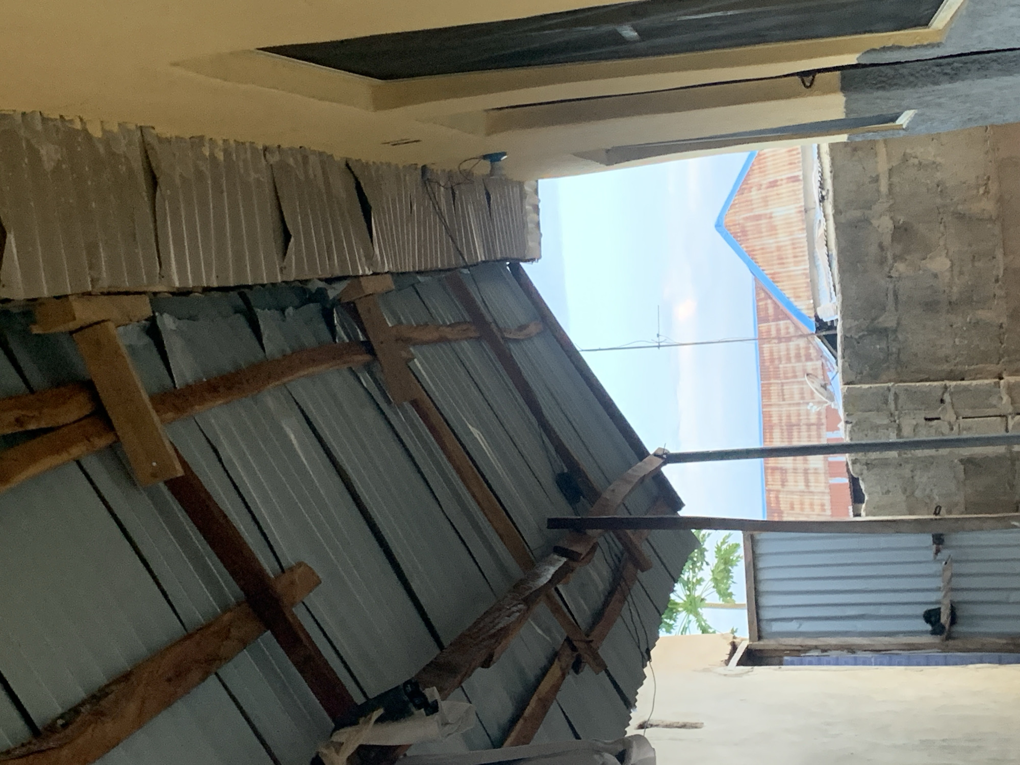 This image, taken in Keffi, Nigeria, depicts an outdoor scene with a focus on mixed architectural elements, viewed from a low angle looking upwards. The primary subject is a makeshift structure in the foreground and midground, composed of corrugated metal sheets (likely zinc or aluminum) held together by a frame of rough-hewn wooden beams and planks. Many of these metal sheets appear weathered, bent, and possibly discolored or rusted. This structure angles upwards from the lower left towards the center.
Above this makeshift construction, on the left side, is a section of a more permanent building with a light beige or yellowish facade and an overhead concrete slab or ceiling. A small, dark-colored device, possibly a security camera or light fixture, is attached beneath this overhang, with visible wiring. A dark rectangular opening, possibly a window or a gap, is also seen on this building.
To the right, a wall built from unrendered concrete blocks (cinder blocks) is visible, suggesting a construction zone or a less finished part of a building. In the background, under a clear, bright blue sky, the roofs of other buildings are discernible. One distant roof features reddish-brown corrugated sheets with a distinctive blue trim along its edge, while another appears to have gray corrugated sheets. An antenna or metal rod extends upwards against the sky.
The scene conveys a sense of utilitarian construction, possibly informal or an ongoing project, in a daytime setting with fair weather. No people or visible text are present.