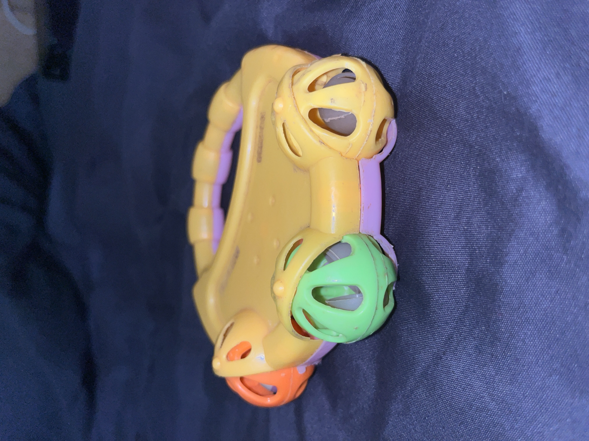 The image shows a yellow and pink plastic toy with orange and green spherical elements. The toy has a curved handle on top and is positioned on a dark blue fabric surface. The toy appears to be a rattle or activity toy, possibly for a baby or toddler, with openings in the spherical parts that may contain internal rattling elements. The lighting is dim, casting shadows and highlighting the texture of the fabric. There is no indication of the location in Abuja, Nigeria; the scene is indoors or in a controlled environment with a neutral background.