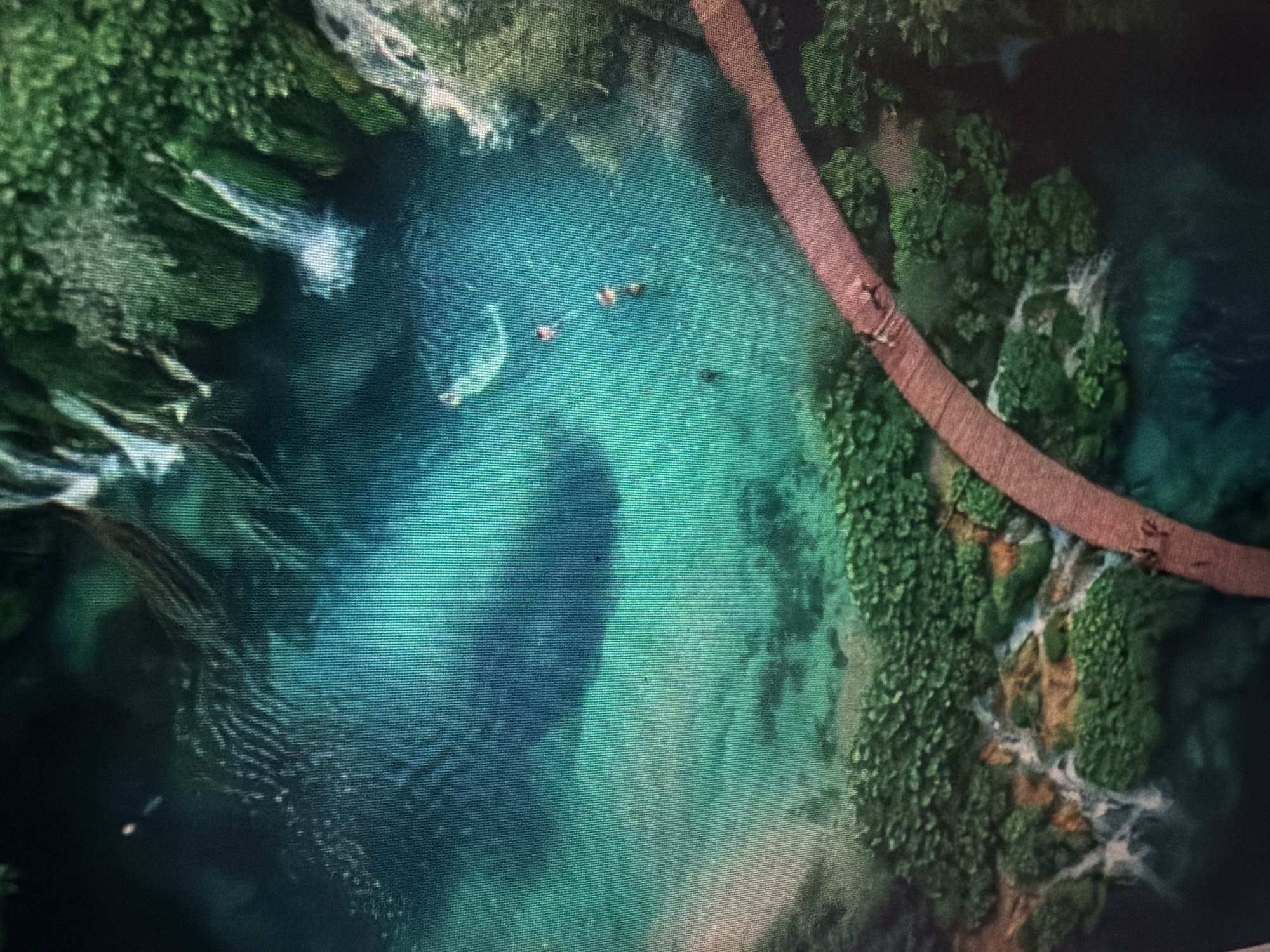 This is an aerial drone shot of a crystal-clear turquoise river flowing through a lush green forest. A rustic, wooden boardwalk curves along the right bank of the river, with several people visible walking on it. In the center of the river, a small group of people are swimming and enjoying the water. The water is shallow in some areas, revealing the sandy riverbed below, while other parts are a deep, inviting blue. The surrounding vegetation is dense and vibrant, suggesting a tropical climate. The lighting suggests it is daytime, with no indication of specific weather conditions. The scene conveys a sense of peaceful recreation and natural beauty. No text is visible in the image.