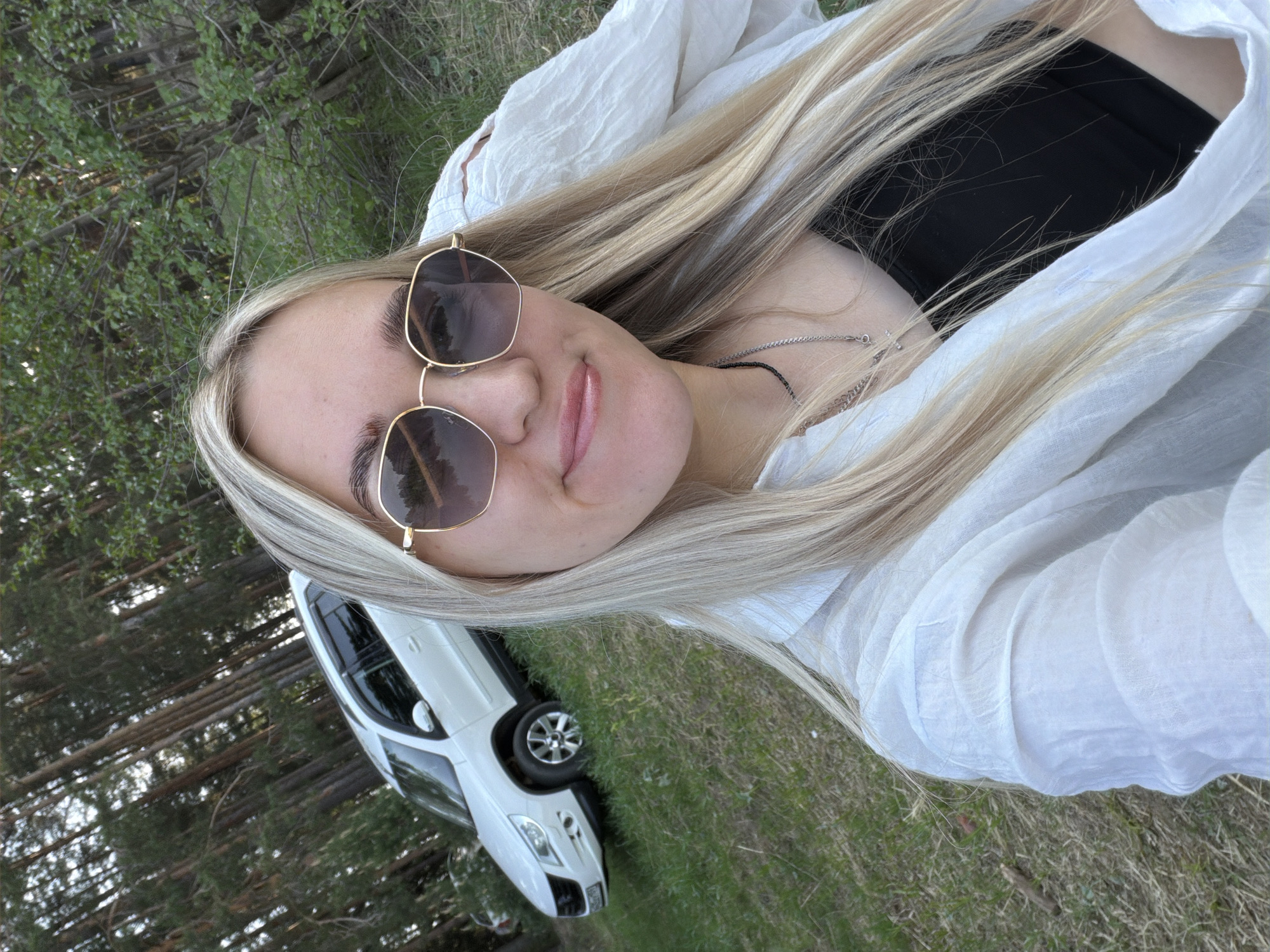 A woman with long blonde hair and sunglasses is taking a selfie outdoors. She is wearing a white shirt over a black top. A white car is partially visible in the background, along with trees and grass. The location is Unknown, Unknown.
