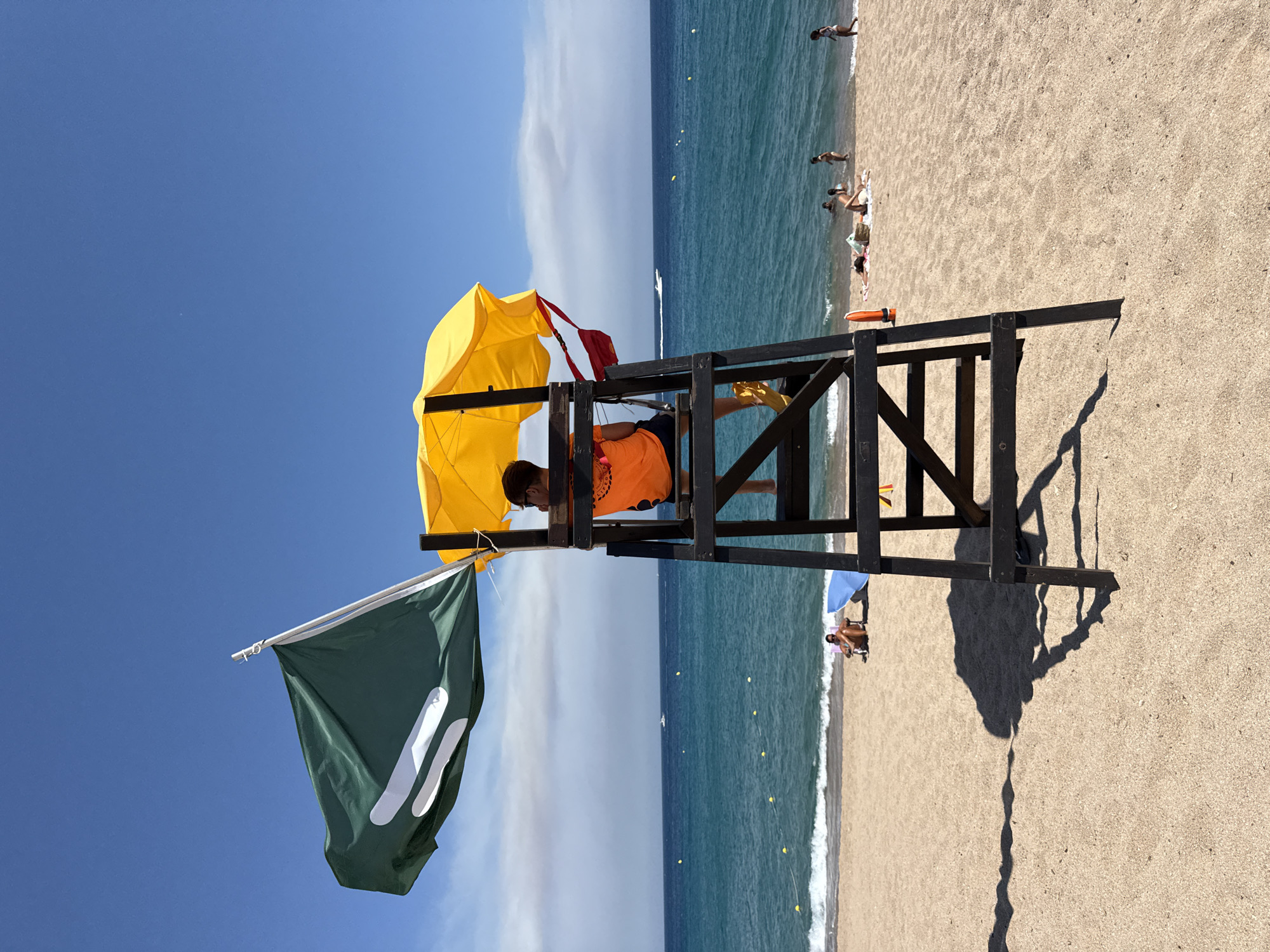 A lifeguard sits on a tall wooden chair on a sandy beach in Blanes, Spain, facing the ocean. The lifeguard wears an orange t-shirt and dark shorts, with yellow shoes. A yellow umbrella is positioned above the lifeguard's head for shade. A green flag with white markings flies from a pole attached to the lifeguard chair. The beach is populated with several people, some sunbathing and others in the shallow water. The sea is calm with small waves, and a boat can be seen in the distance. The sky is clear and blue, with some wispy clouds on the horizon. The sun casts long shadows from the lifeguard chair onto the sand. The scene depicts a typical summer day at the beach.