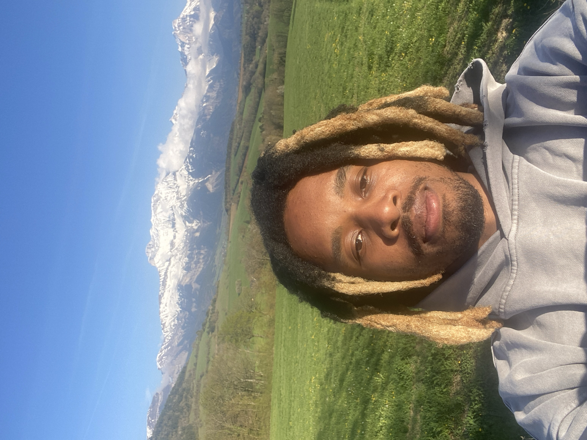 A medium shot depicts a person with dark skin, dark facial hair, and blond-tipped dreadlocks. They are wearing a gray hooded sweatshirt. The background shows a bright blue sky and a grassy field leading to a mountain range partially covered in snow. The photo was taken during daylight. Location: Cornillon-en-Trièves, France.