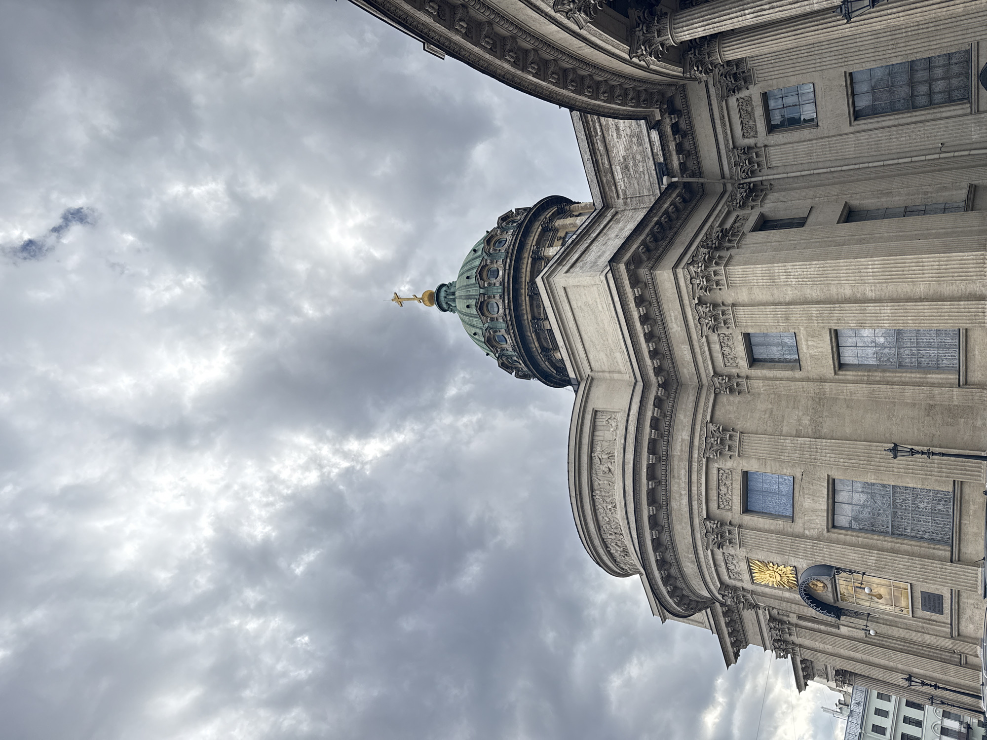 The image depicts a partial view of the Kazan Cathedral in Saint Petersburg, Russia, taken from a low angle looking upwards. The cathedral's green dome with a golden cross is visible against a backdrop of overcast skies. The cathedral's facade is ornate, with multiple levels of detailing, columns, and windows. A golden emblem is visible on the side of the building, along with a plaque and lighting fixture. Other buildings and structures are visible in the very bottom right of the image. The sky is filled with gray clouds.