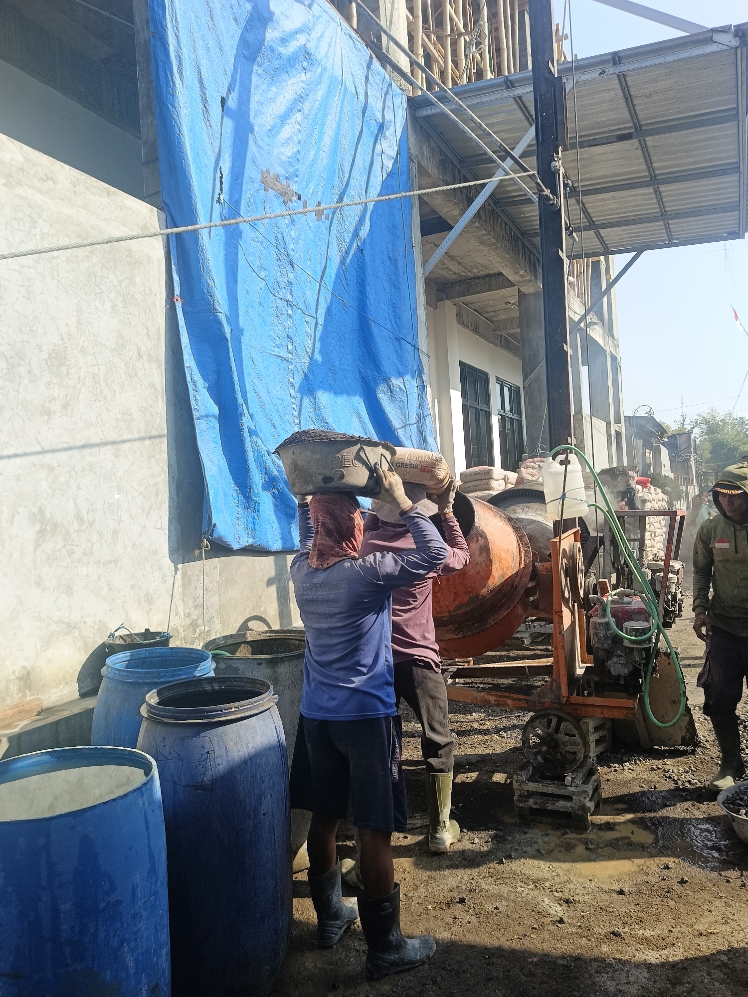 In Gedangan, Indonesia, a construction site is captured in a medium shot, bathed in bright daylight. The scene centers on two men wearing work attire and head coverings, actively engaged in construction activities. One man, positioned on the left, balances a large basin filled with soil on his head, his back towards the viewer, and is assisted by another man. They are near an industrial cement mixer, an orange and metal contraption with a green hose attached to a white jug. Several large blue plastic barrels and stacks of cement bags are visible, suggesting ongoing construction work. In the background, unfinished building structures with exposed bamboo scaffolding rise against a clear sky. Another man, wearing a hooded jacket, stands to the right, observing the activity. The ground is uneven, covered with dirt, gravel, and puddles of water, indicating a potentially muddy environment.