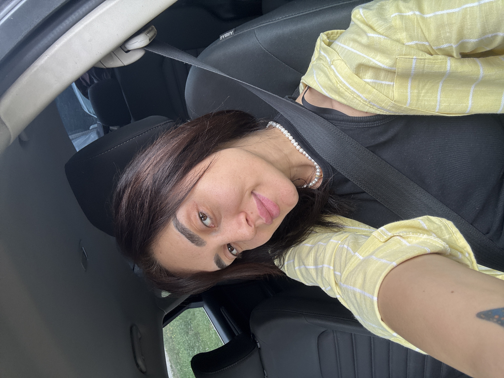 This is a selfie taken by a woman inside a car. She is wearing a black top and a light yellow and white striped open shirt. A pearl necklace is visible around her neck. She has dark brown hair and is looking towards the camera with a slight smile. The car interior is visible, including a black seat, seatbelt, and parts of the car's interior trim. The background outside the car appears to be a green, natural landscape, suggesting the car is in motion or parked in a rural or suburban area. The lighting suggests it might be daytime. The visible text in the image reads "AIRBAG". The image appears to be geotagged to Tbilisi, Georgia.