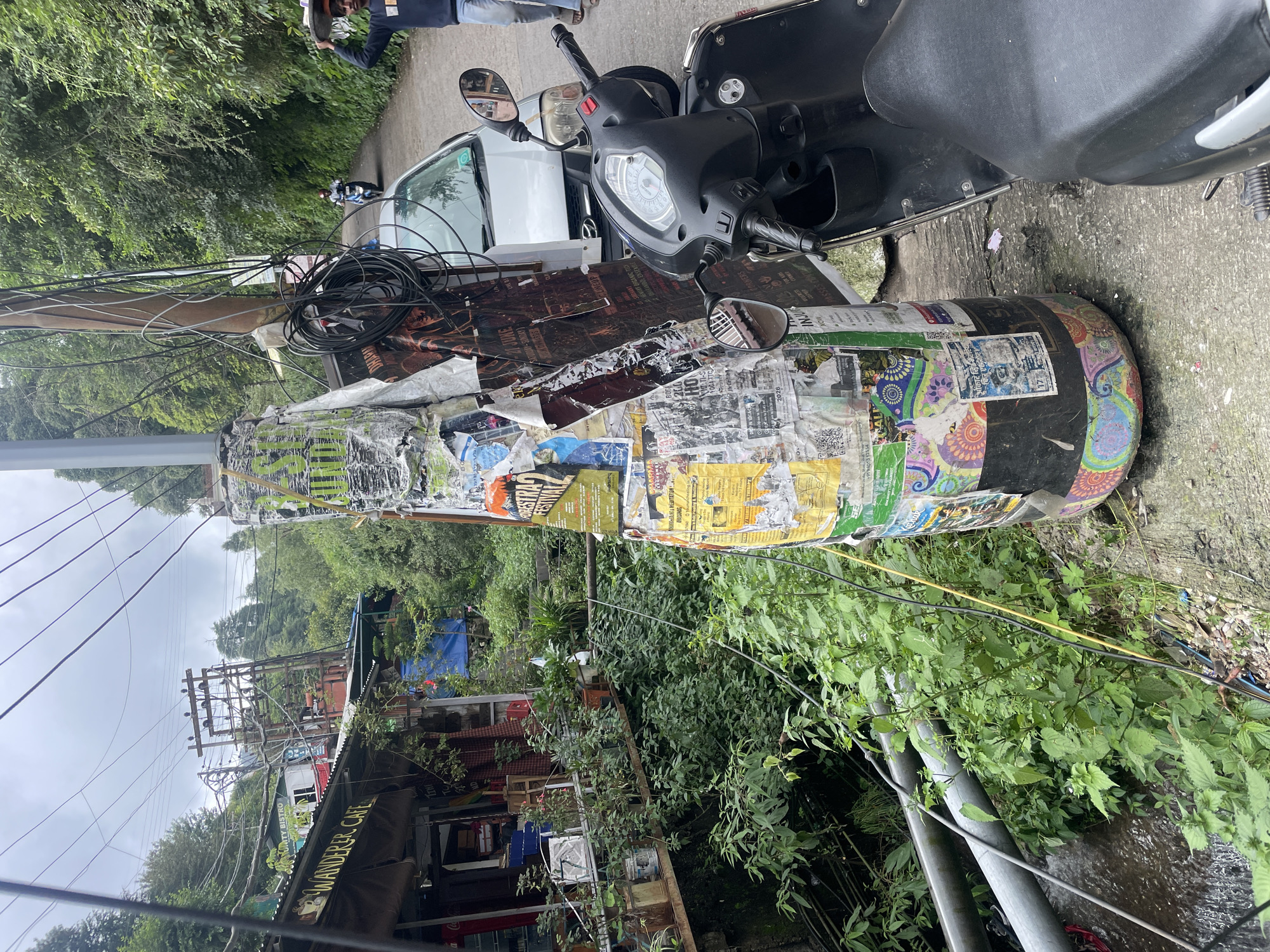 The image depicts a street scene in Bhagsu, India, characterized by lush greenery and a somewhat cluttered environment. In the foreground, a large, cylindrical structure, resembling a giant bottle, is covered in various posters and colorful artwork. Electrical wires and cables are prominently visible, draped across utility poles and the bottle structure, suggesting a dense network of electrical infrastructure.
To the left of the bottle, a building with a corrugated metal roof houses "Wanderer Cafe," with tables and chairs visible through the open facade, and plants adorning its exterior. The cafe appears to be built on an elevated platform, with a stream or drainage channel running beneath it, flanked by thick vegetation.
On the right side of the frame, a black scooter is parked on the paved road. A silver car is also visible further down the road. In the background, a man is walking away from the camera, carrying something on his head. The road winds uphill, disappearing into a densely wooded area covered with trees. The sky is overcast, contributing to a subdued lighting. The overall impression is of a vibrant, yet somewhat chaotic, urban environment integrated with nature.