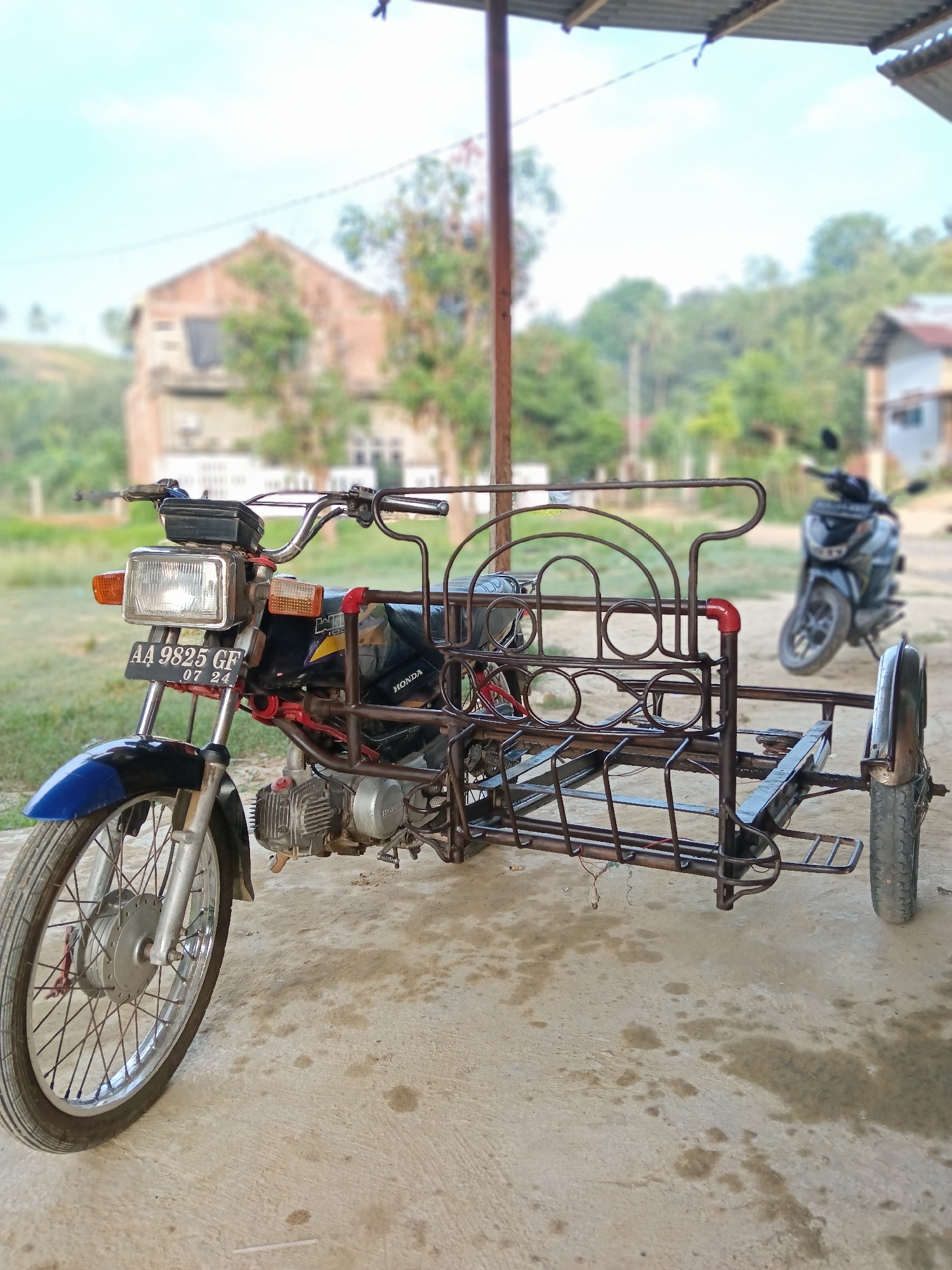 The image depicts a motorcycle with a sidecar parked on a concrete surface, possibly a patio or driveway, in what appears to be a semi-rural setting. The motorcycle is older, with a square headlight, blue fender, and a license plate reading "AA 9825 GF 07 24". The sidecar is constructed of metal tubing with circular cutouts and a small wheel. In the background, there is a blurred building, possibly a house or small business, and another motorcycle parked further away. The environment includes trees and a slightly hilly landscape under a bright sky. Considering the license plate style and general scenery, the location is in Indonesia, and given the prompt information, it is in Lhokseumawe.